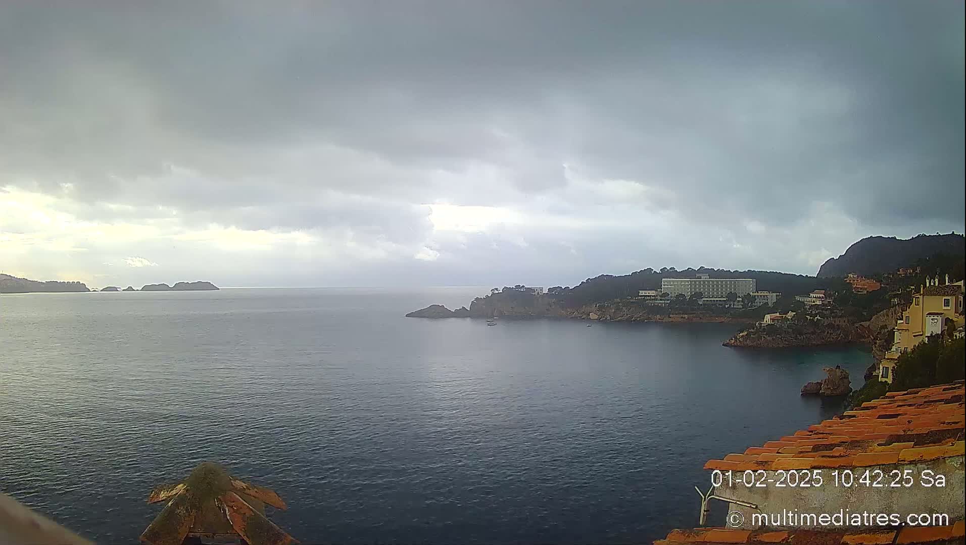 A partly cloudy, overcast day shows a coastal view of a calm bay with distant islands, a hotel, and buildings on a rocky shoreline.