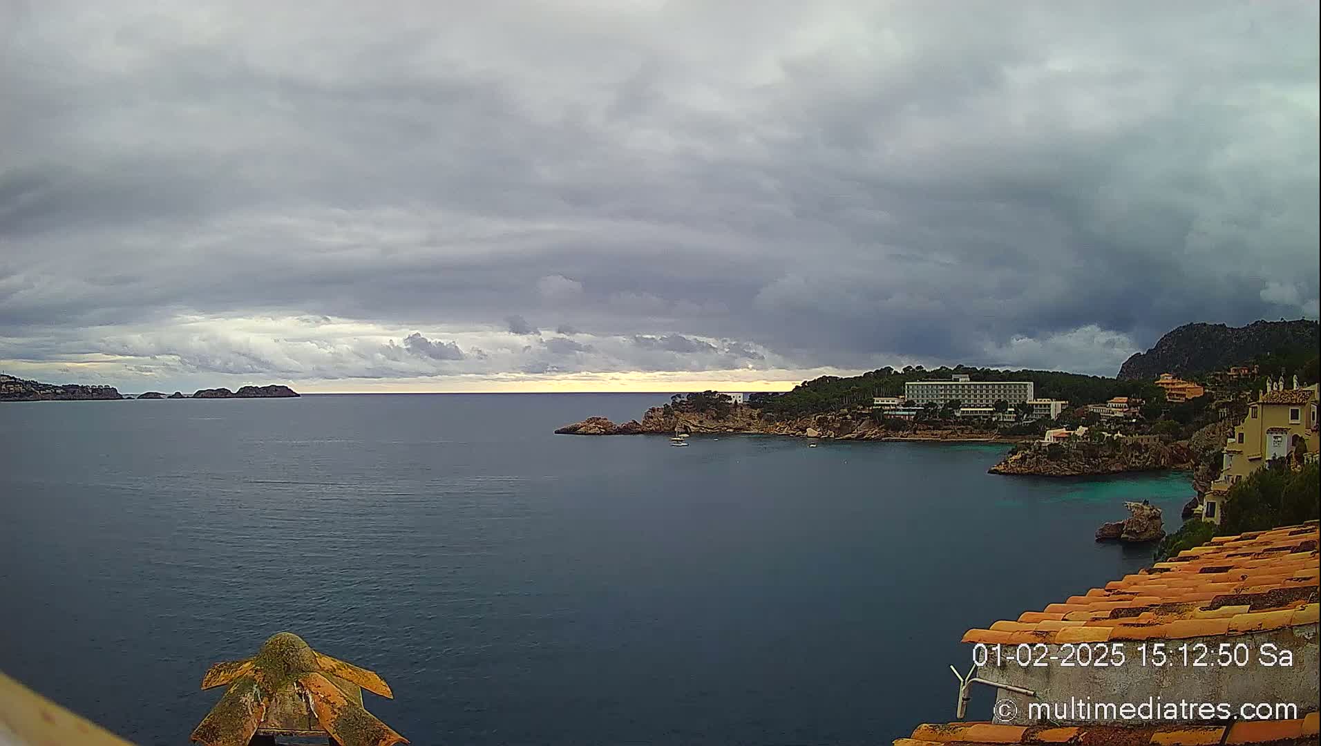 A coastal scene under a cloudy sky shows a calm body of water with a hotel and buildings on a rocky shore, and small islands in the distance.