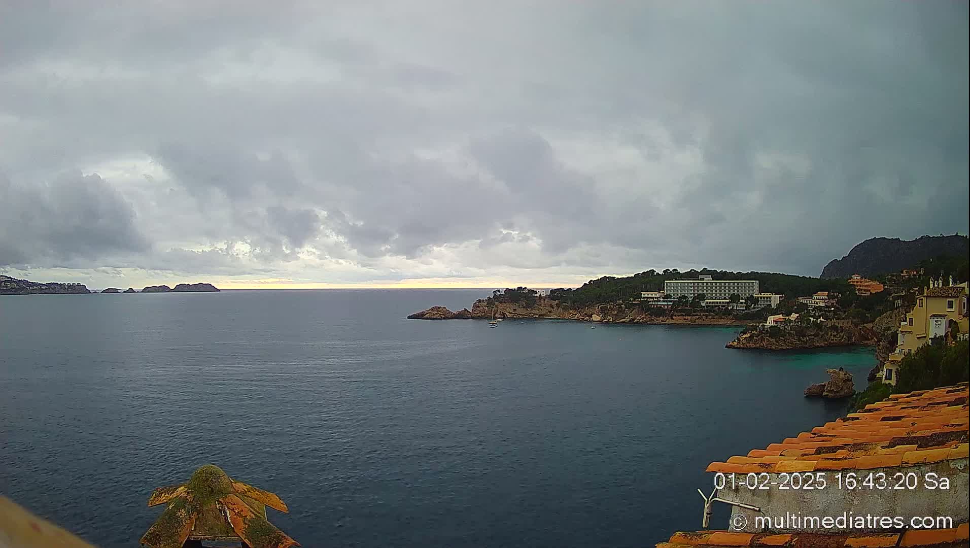 A coastal scene under a cloudy sky shows a calm sea with distant islands and a shoreline featuring buildings and vegetation.