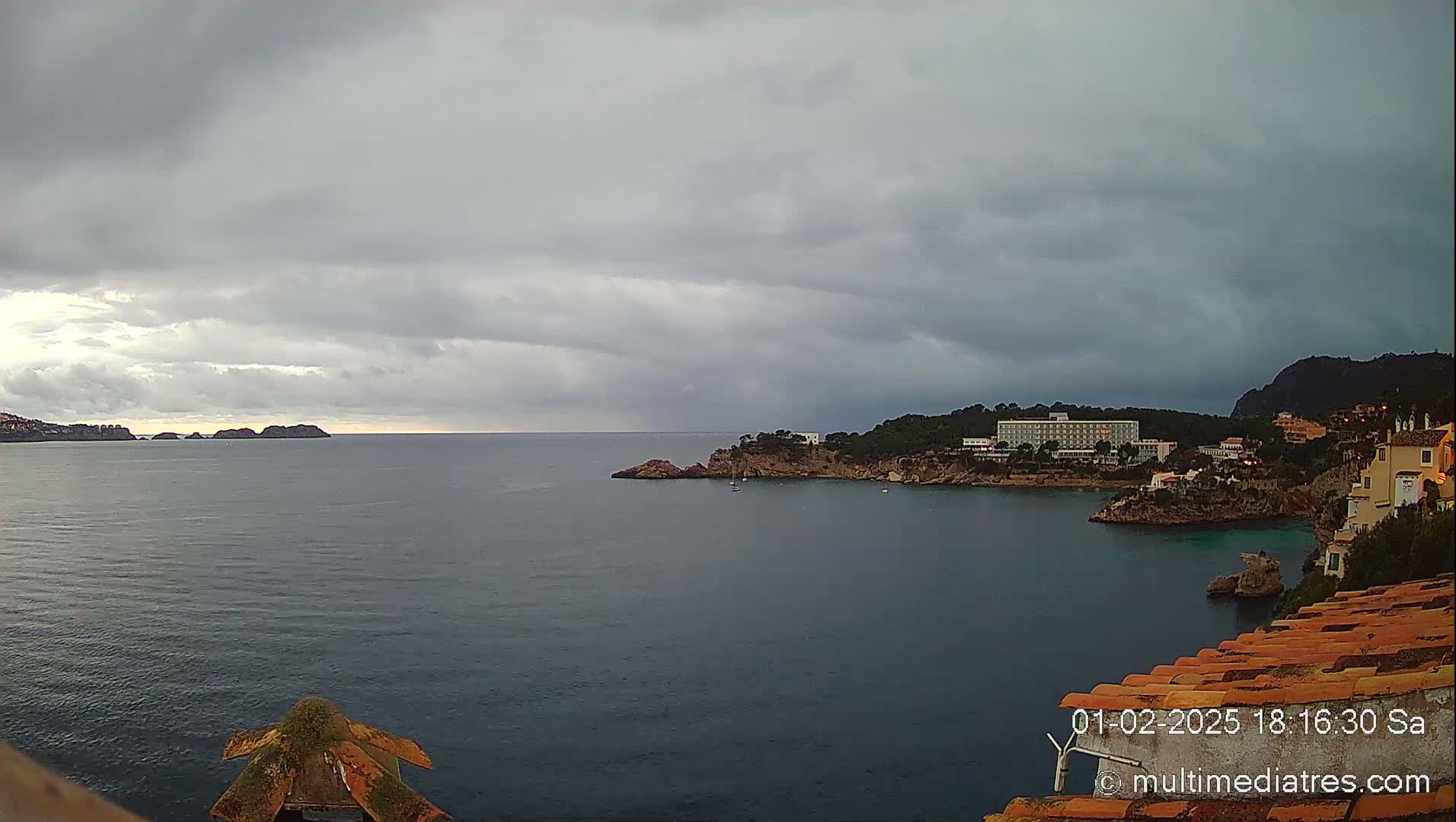 A coastal scene under a cloudy sky shows a calm bay with a hotel and buildings on a rocky shoreline, and distant islands.