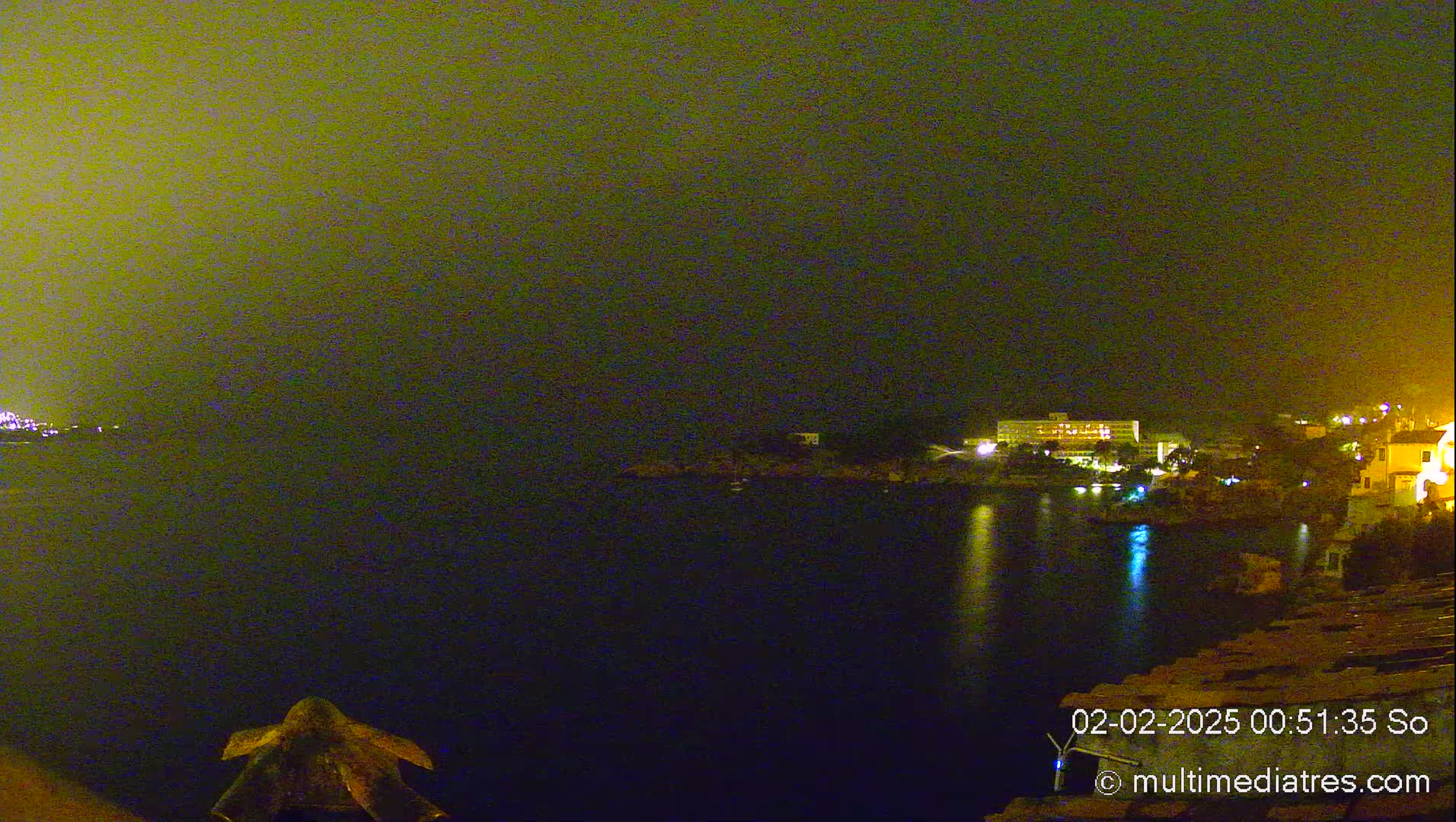 A nighttime view of a calm body of water with illuminated buildings and vegetation on the far shore under a hazy, yellowish sky.