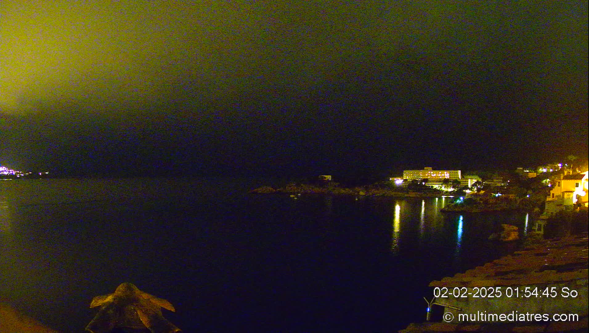 A nighttime coastal scene shows a dark body of water reflecting lights from buildings on the opposite shore under a hazy, dark yellow sky.