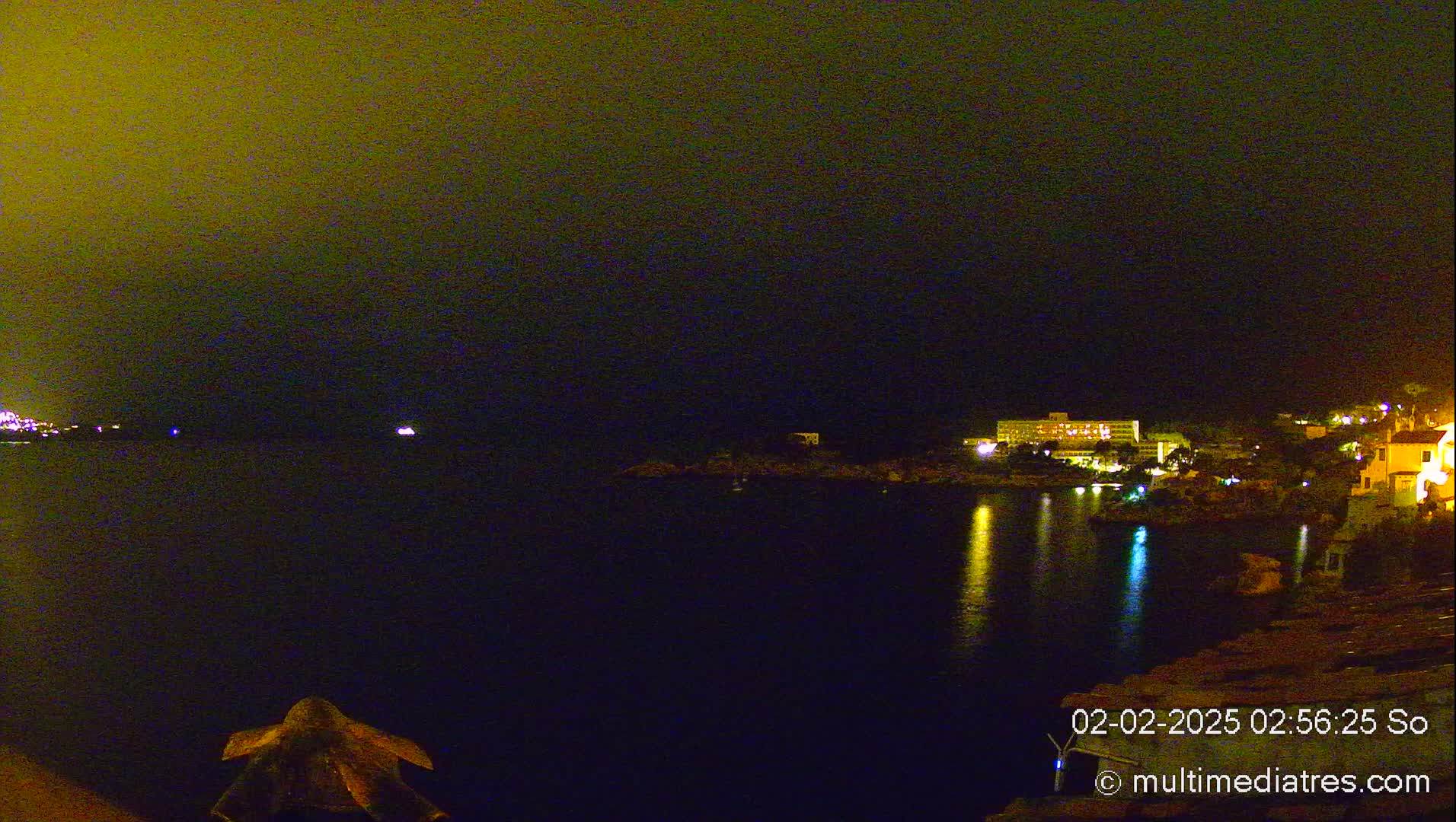 A nighttime view of a dark body of water reflecting the lights of buildings on a shore under a hazy, yellow-tinged sky.