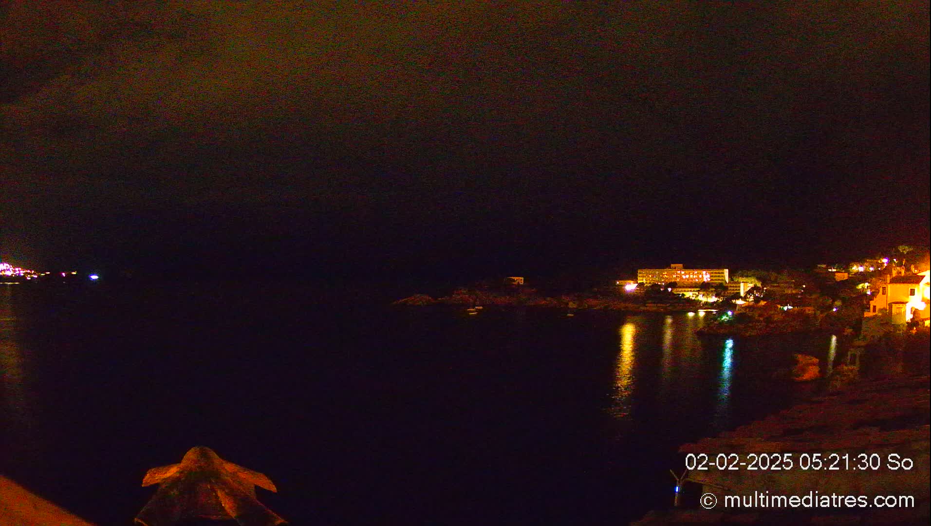 A nighttime view of a dark body of water reflecting the lights of buildings on a distant shore under a cloudy sky.