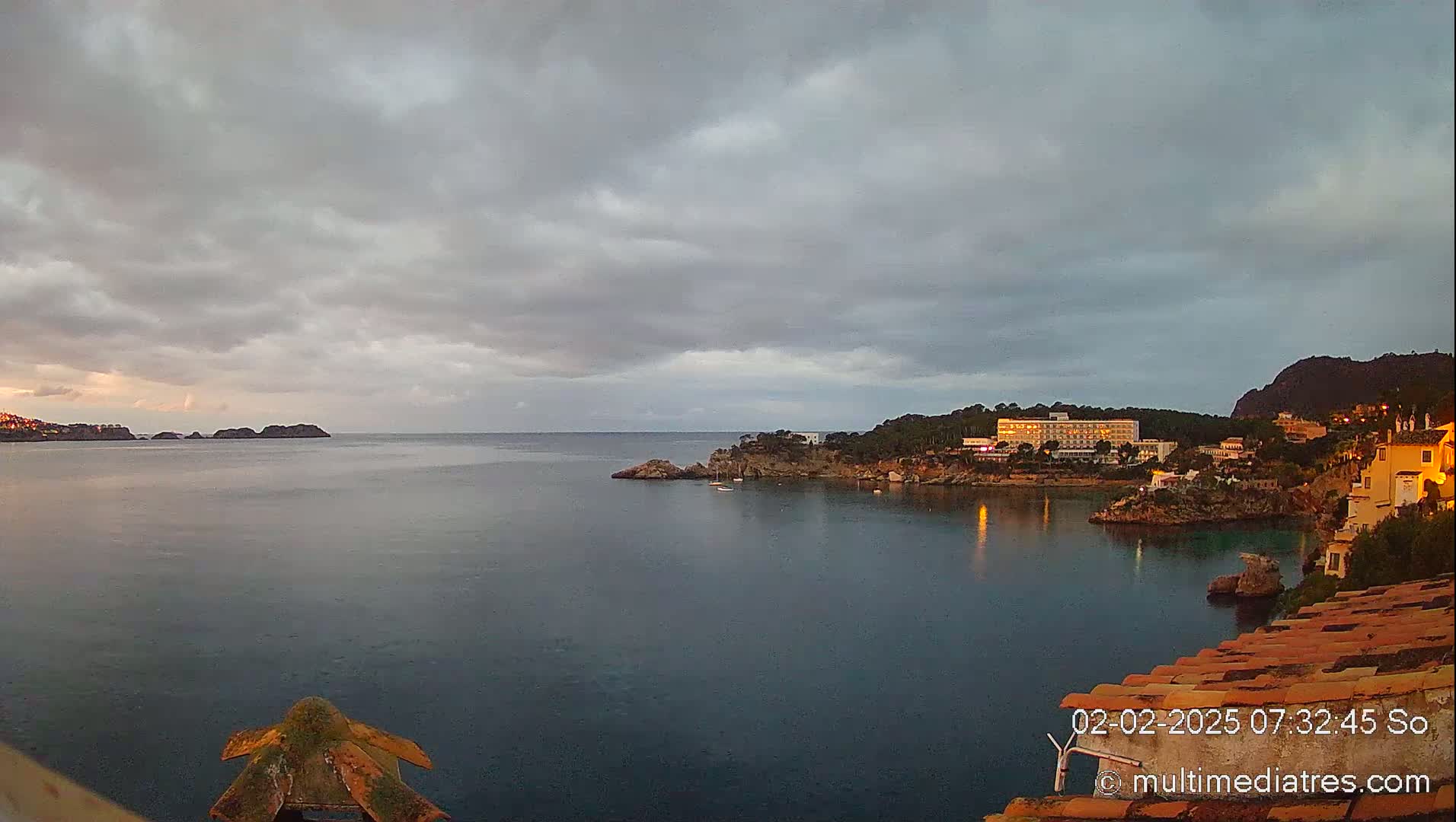 A calm, dark-blue bay is nestled between rocky hills under a cloudy, overcast sky, with a large hotel visible on one shore.
