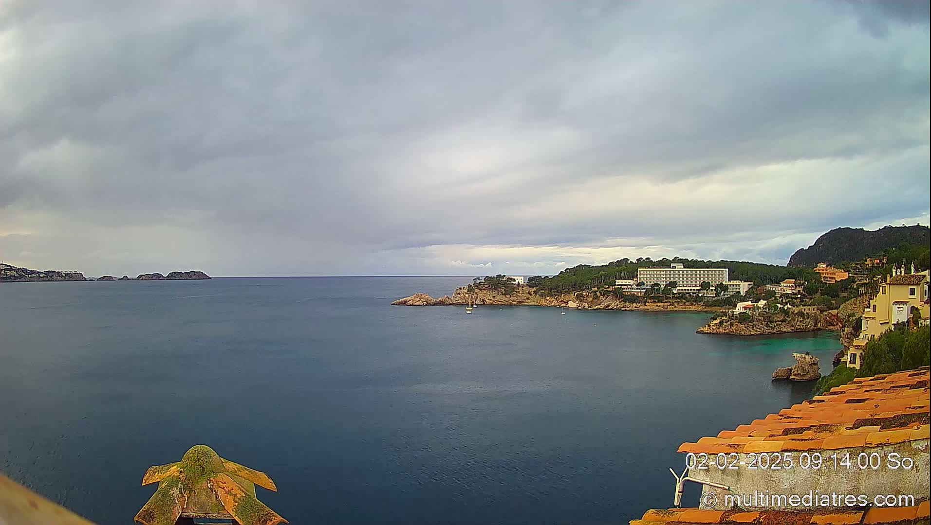A coastal scene under a cloudy sky shows a calm bay with a hotel and buildings on a rocky shore, and a distant landmass.