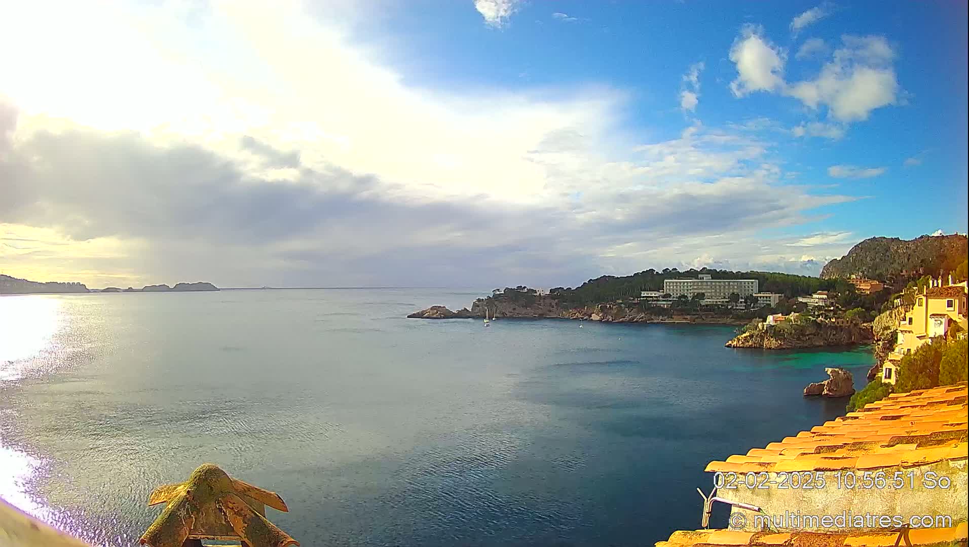 A partly cloudy day reveals a coastal scene with a calm sea, a large hotel, and several buildings situated on a rocky hillside.