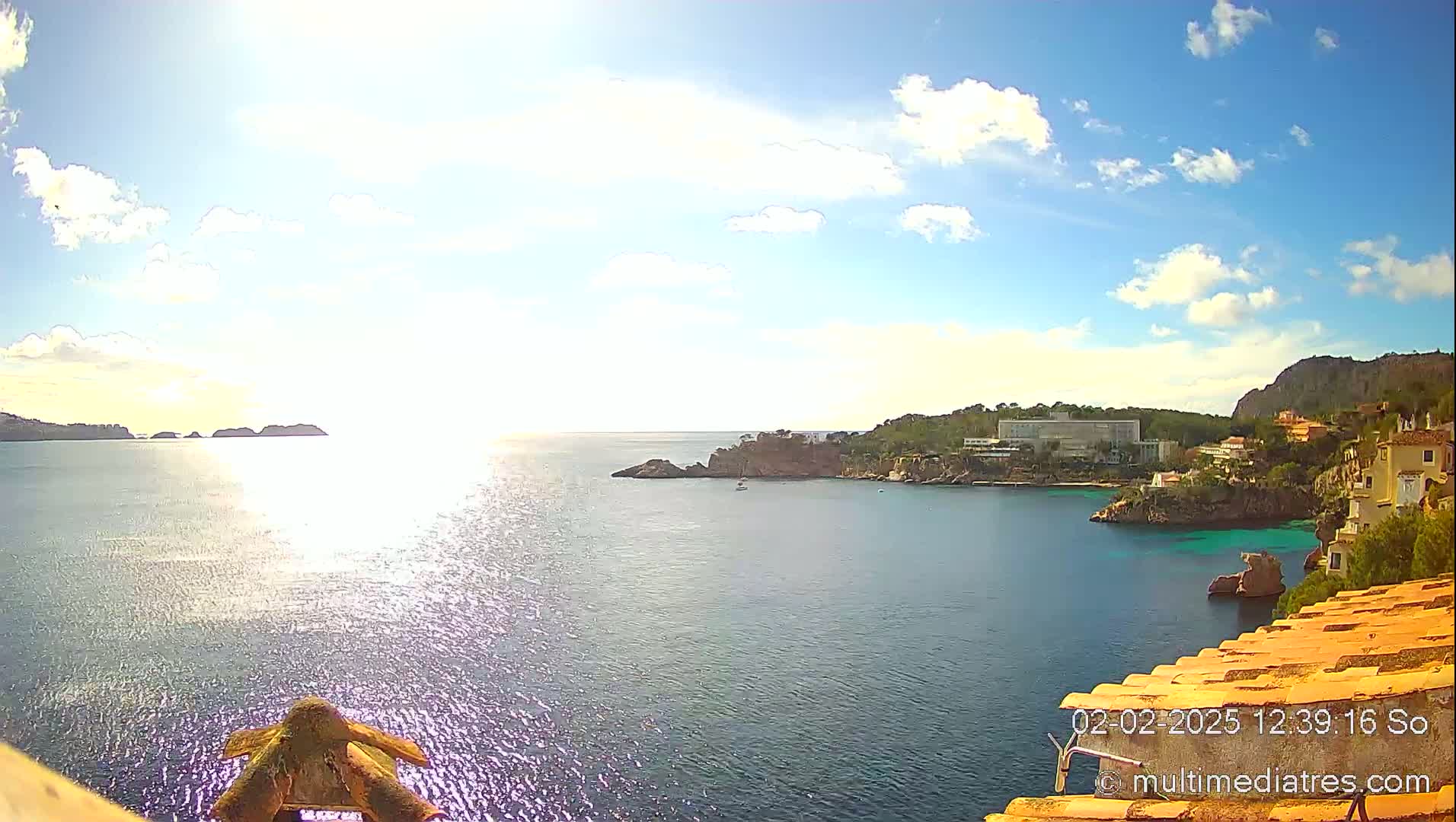 A bright, sunny day with scattered clouds shows a tranquil bay with a hotel and residential buildings along the coastline, small islands in the distance, and sparkling water.