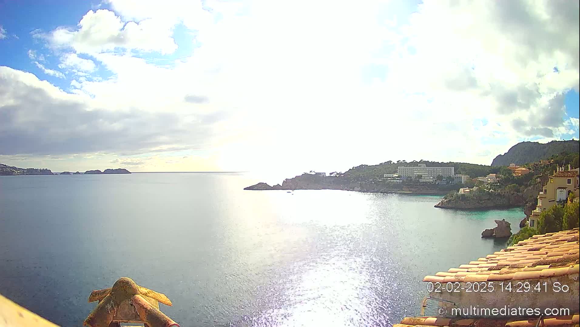 A partly cloudy sunny day overlooking a calm bay with buildings and rocky coastlines.