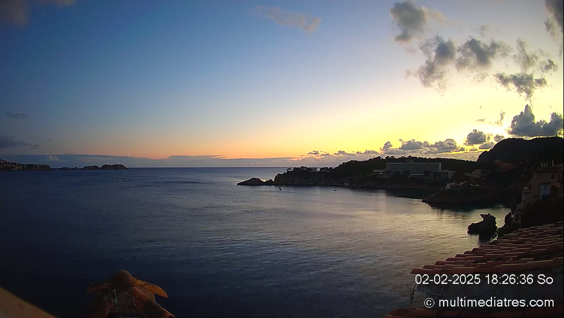 A coastal sunset scene shows a calm sea with dark silhouettes of land, buildings, and a few small boats, under a partly cloudy sky with yellow and orange hues near the horizon.