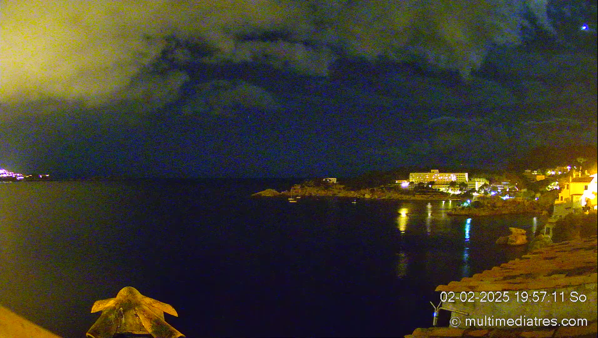 A nighttime coastal scene shows a dark body of water reflecting the lights of buildings on the shore under a partly cloudy sky.