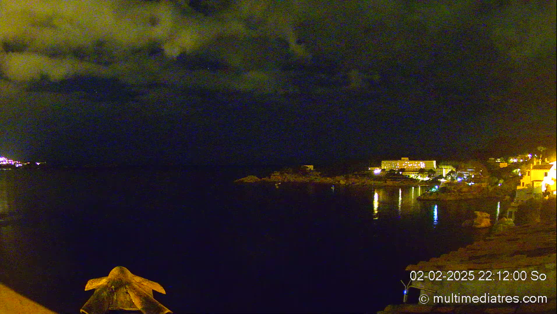 A nighttime view of a calm body of water reflecting the lights of buildings on the opposite shore under a cloudy sky.