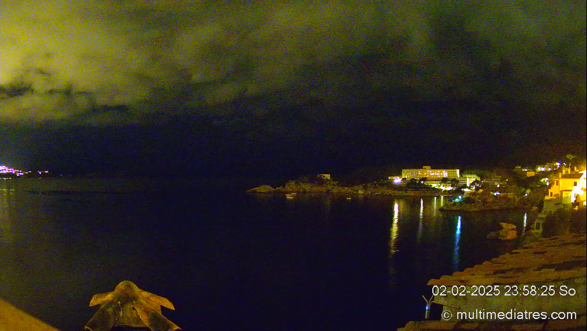 A nighttime view of a calm body of water reflecting the lights of buildings on a distant shore under a cloudy sky.