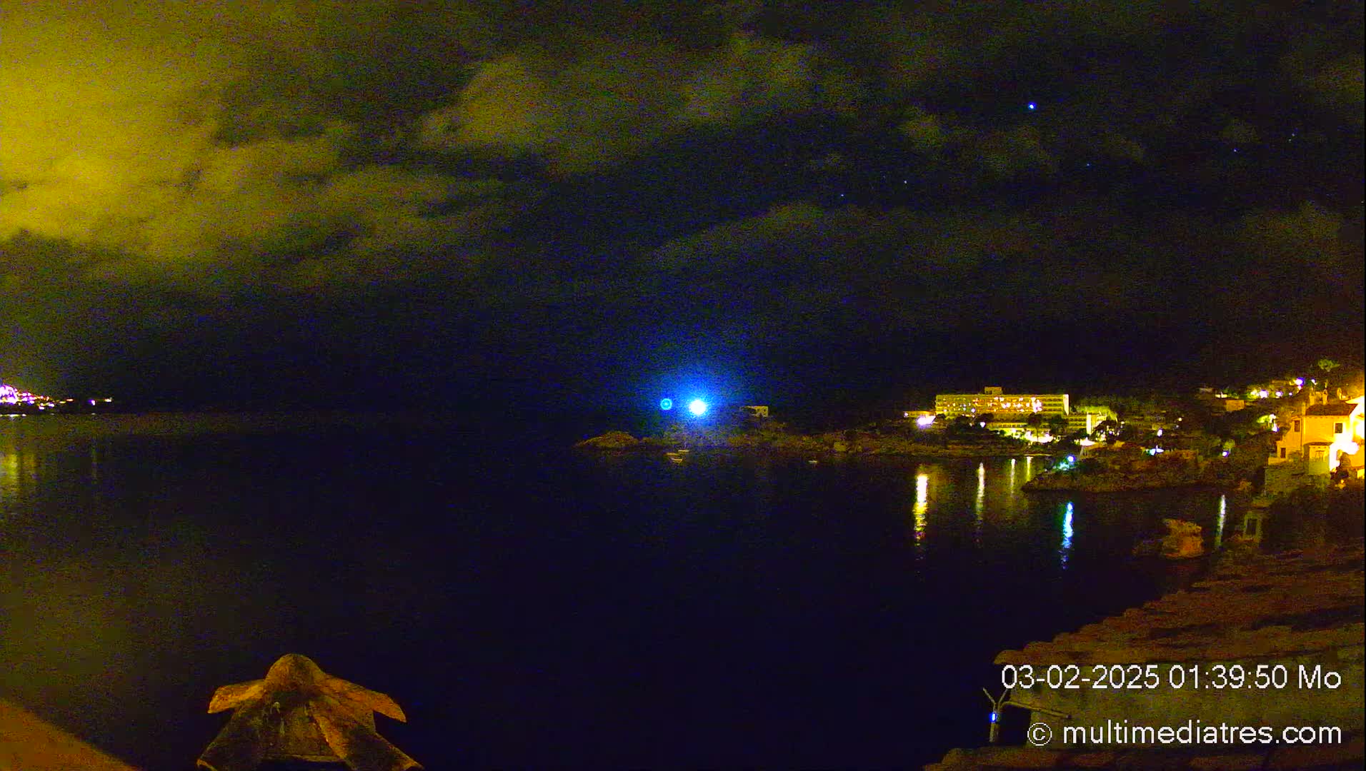 A nighttime view of a calm body of water reflecting the lights of buildings on the opposite shore under a partly cloudy sky.