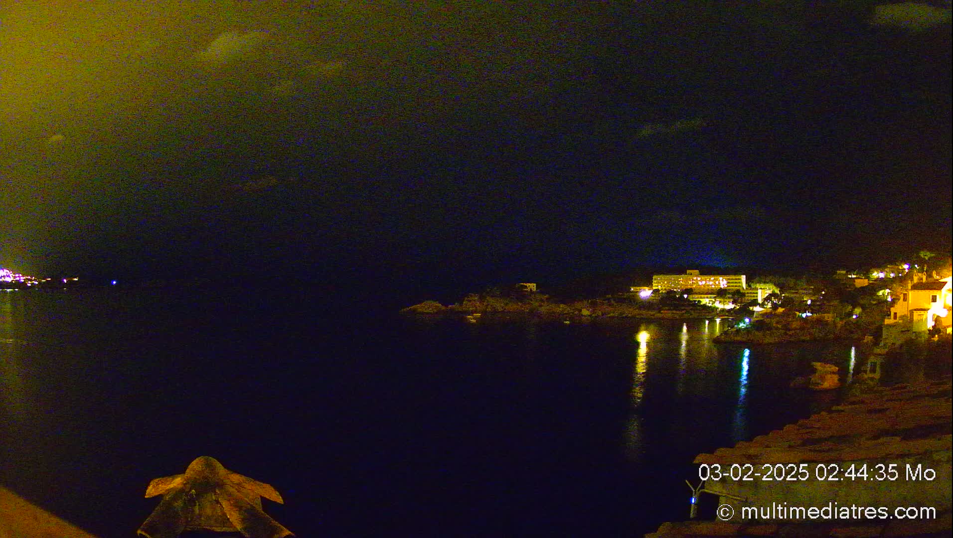 A nighttime view of a calm body of water reflecting the lights of a coastal town under a dark, cloudy sky.