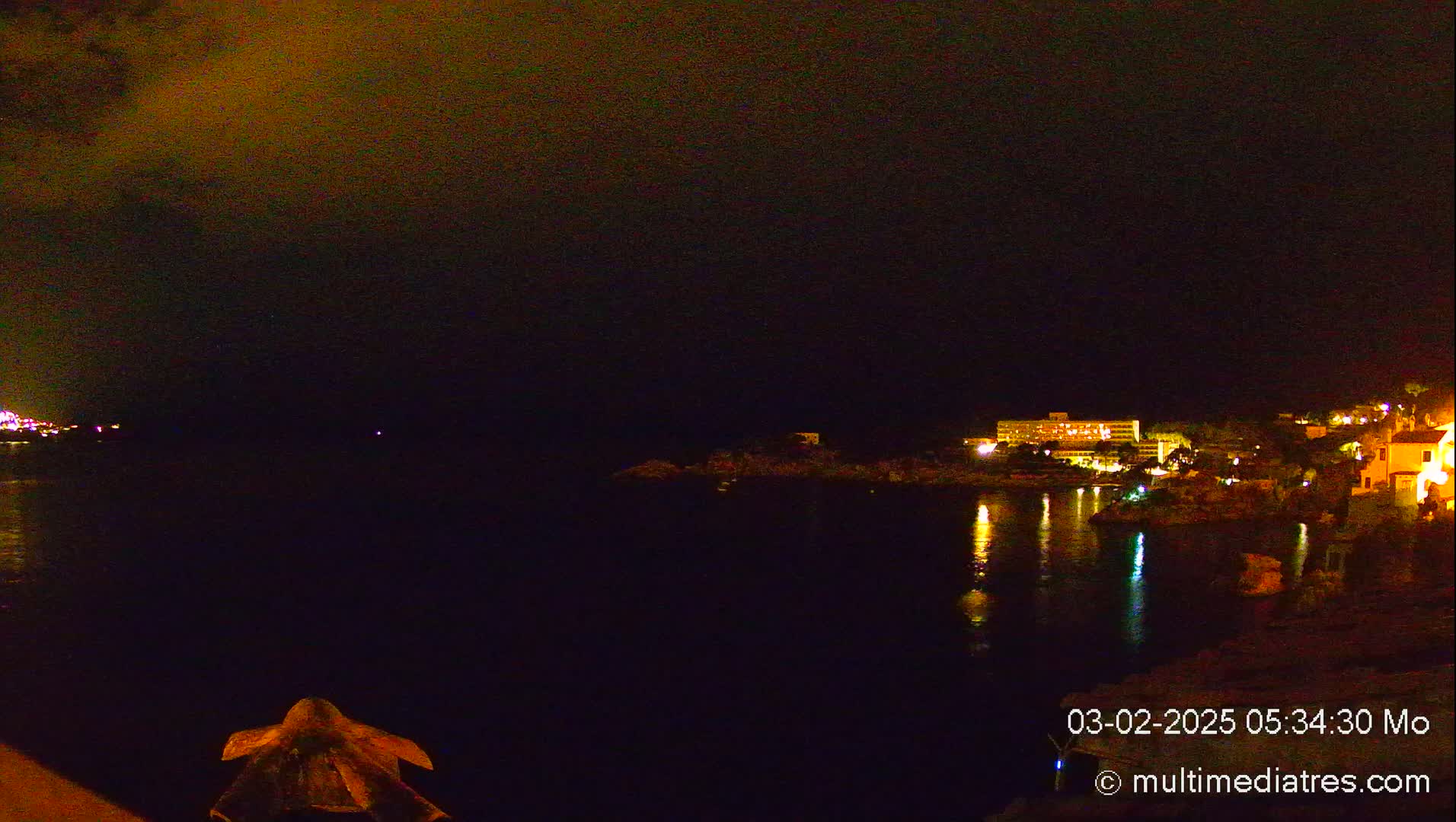 A nighttime view of a calm body of water reflecting the lights of buildings on the opposite shore under a dark, cloudy sky.