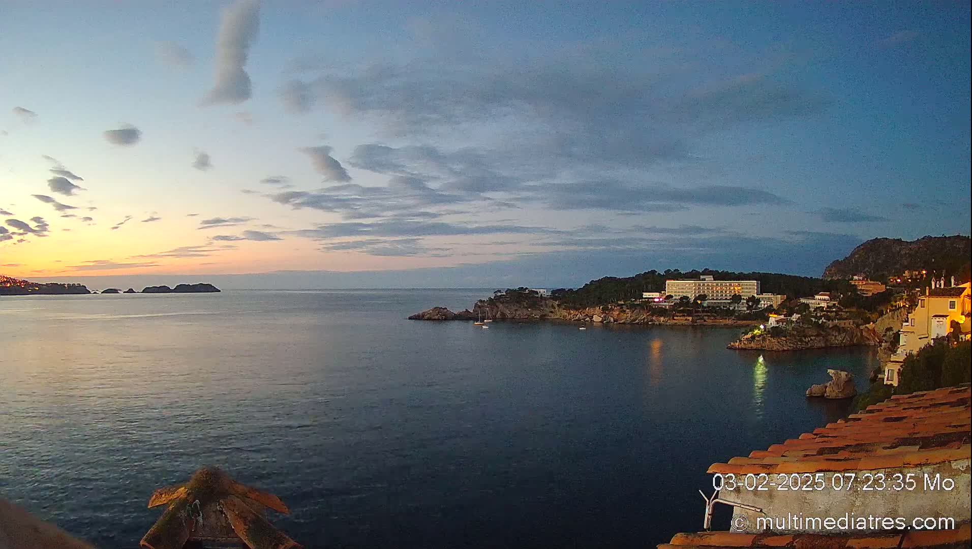 A tranquil coastal scene at sunset features a calm sea, small islands in the distance, a shoreline with buildings, and a partly cloudy sky.