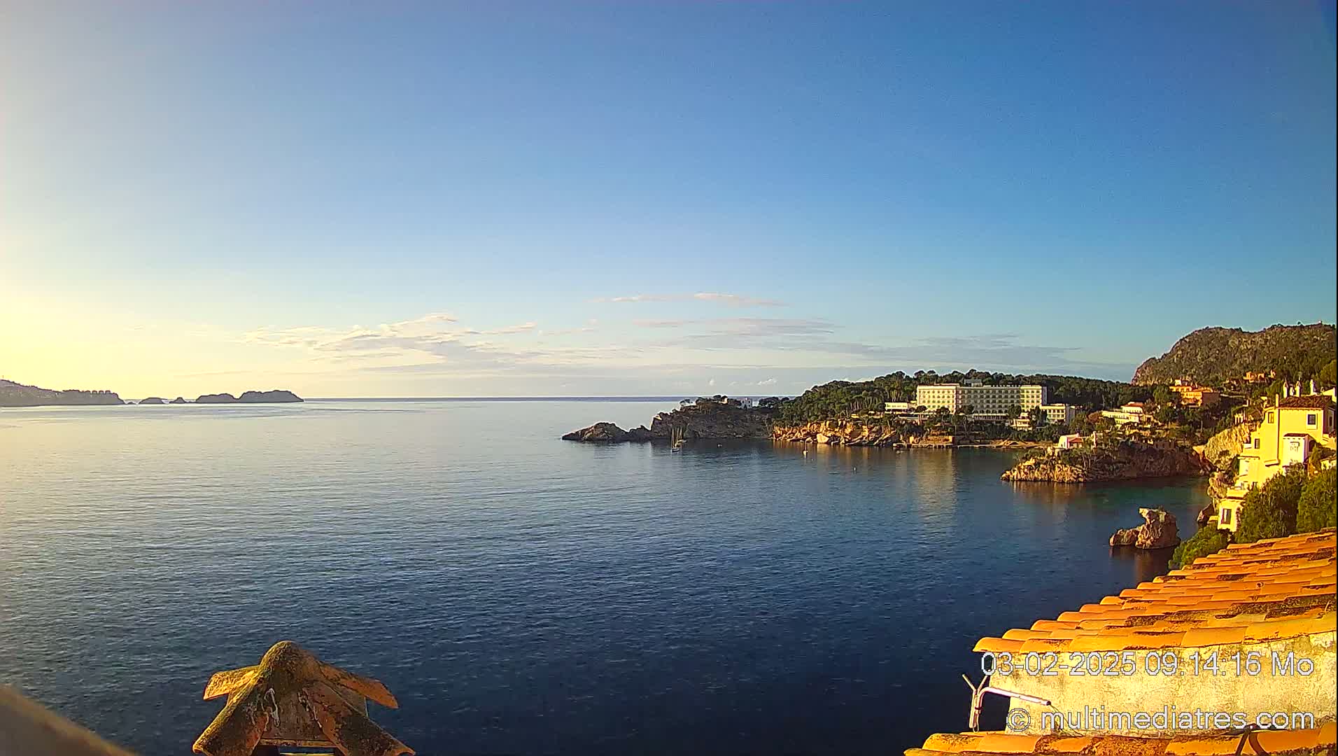 A calm, clear morning shows a coastal view of a hotel and buildings nestled on a rocky hillside overlooking a tranquil bay with small islands in the distance.