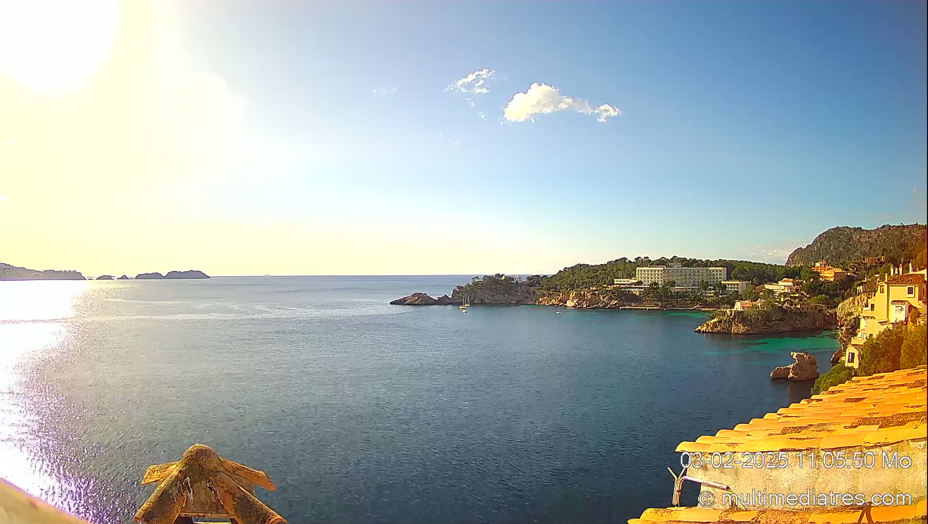 A sunny, clear day reveals a calm ocean cove with a hotel and residential buildings nestled along the rocky coastline, some small boats visible in the water.
