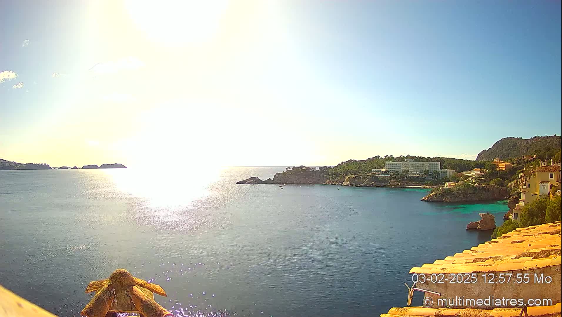 A bright, sunny day overlooks a calm bay with a coastline featuring buildings and lush greenery, small islands dot the horizon.