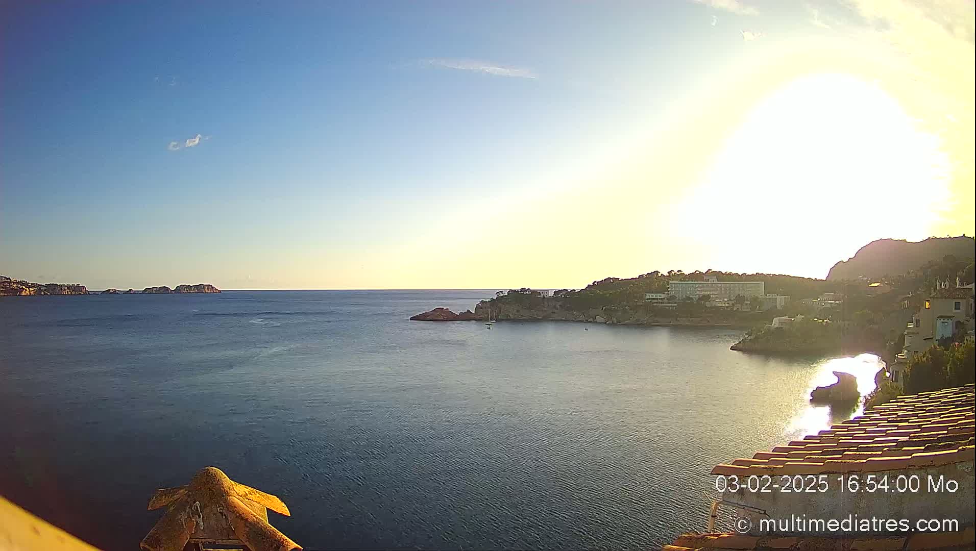A coastal scene at sunset shows a calm sea with rocky outcrops and buildings along the shore under a mostly clear sky.