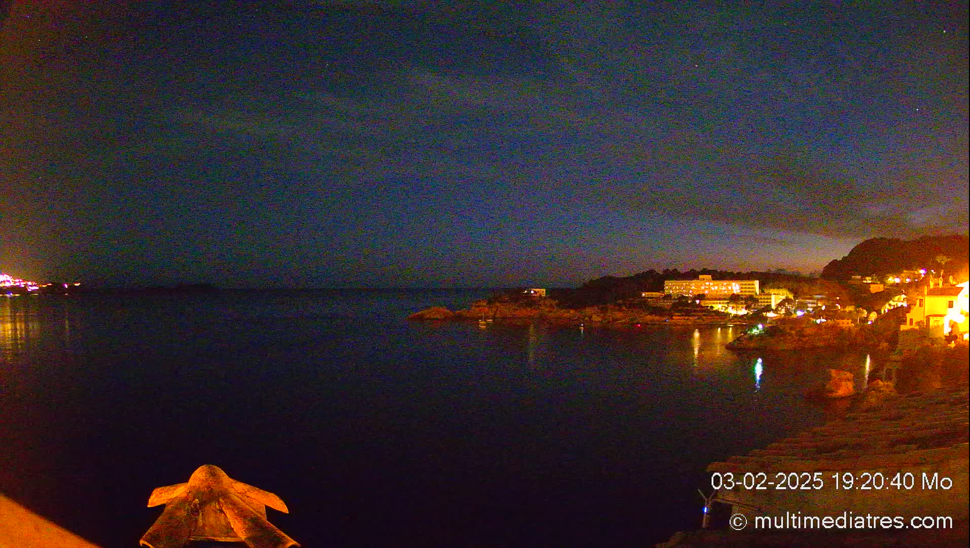 A nighttime view of a calm, dark body of water with illuminated buildings and hills on the far shore under a mostly clear, dark sky.