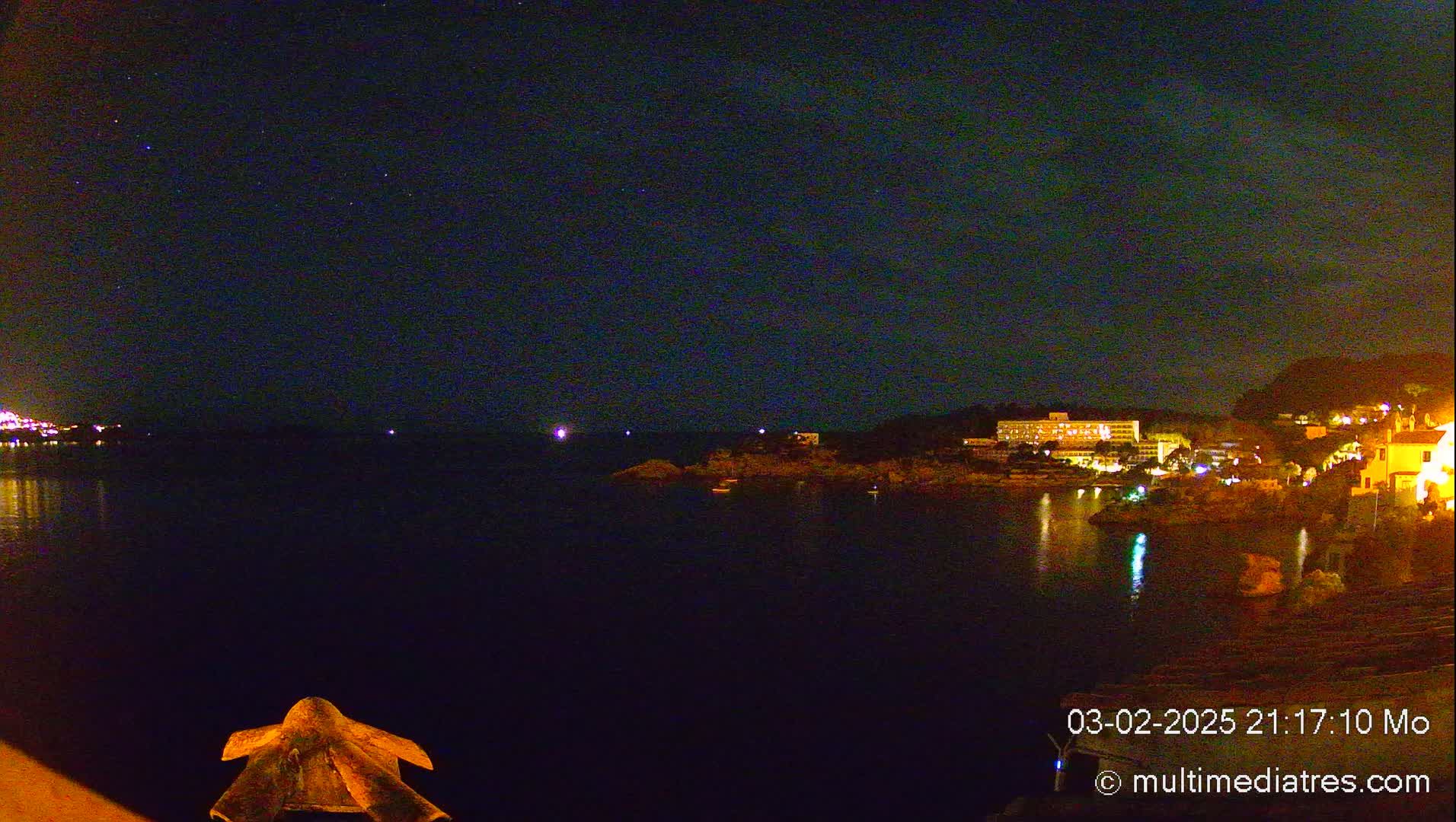 A nighttime view of a dark body of water with a brightly lit shoreline featuring buildings and a starry sky.