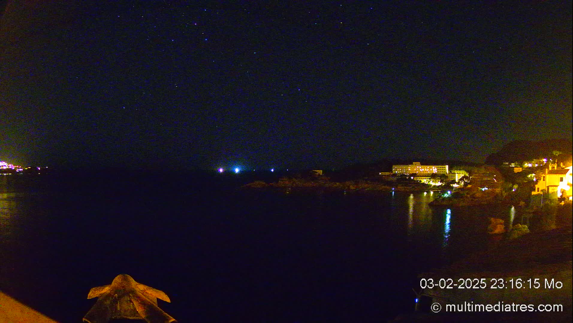 A night scene shows a dark body of water reflecting the lights of buildings on the opposite shore under a clear, starry sky.