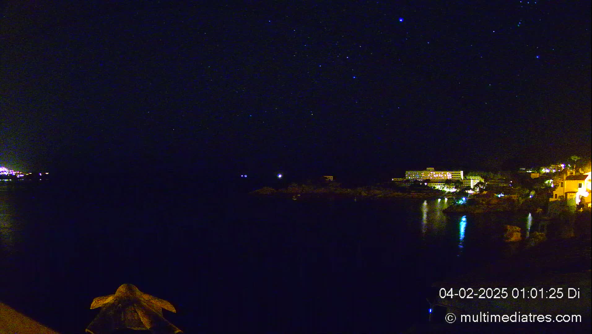 A nighttime view of a calm body of water reflecting the lights of buildings on the shore under a starry sky.