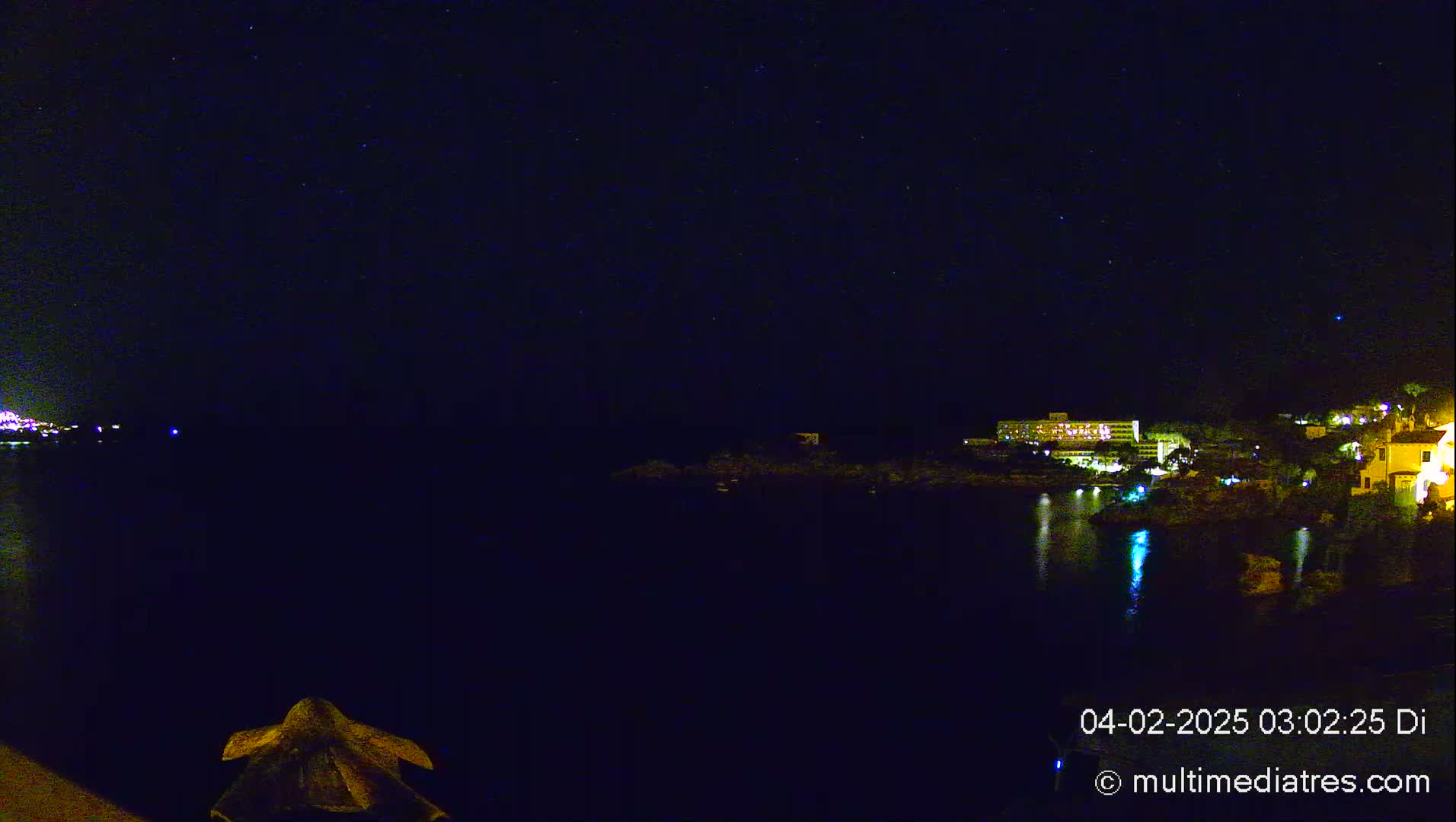 A nighttime view of a calm body of water reflecting the lights of buildings on the shore under a clear, starry sky.