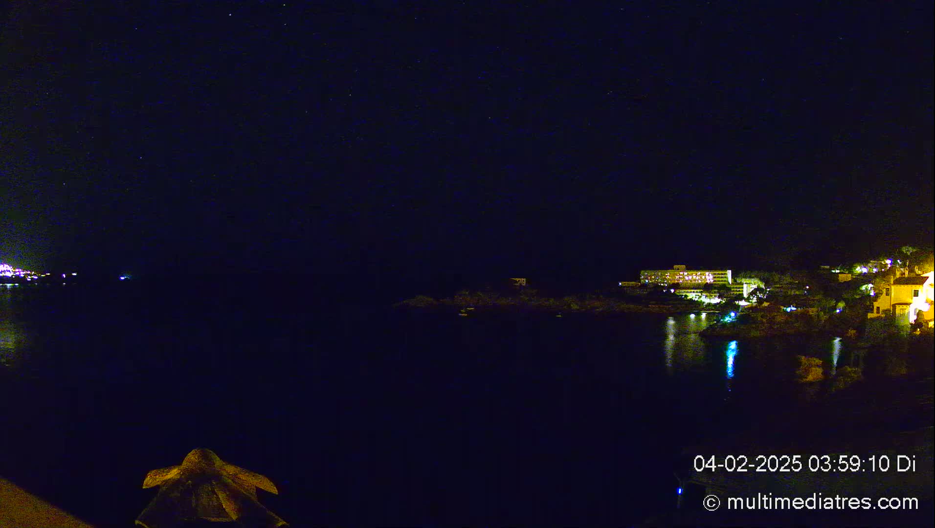 A nighttime view of a calm body of water reflecting the lights of buildings on a distant shore under a starry sky.