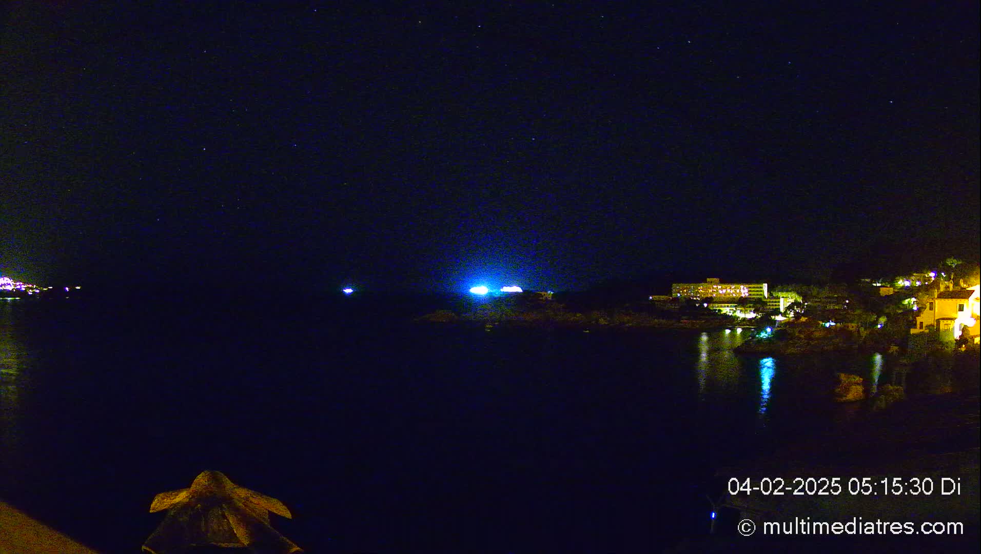 A nighttime view of a calm body of water reflecting the lights of buildings on a shoreline under a clear, starlit sky.