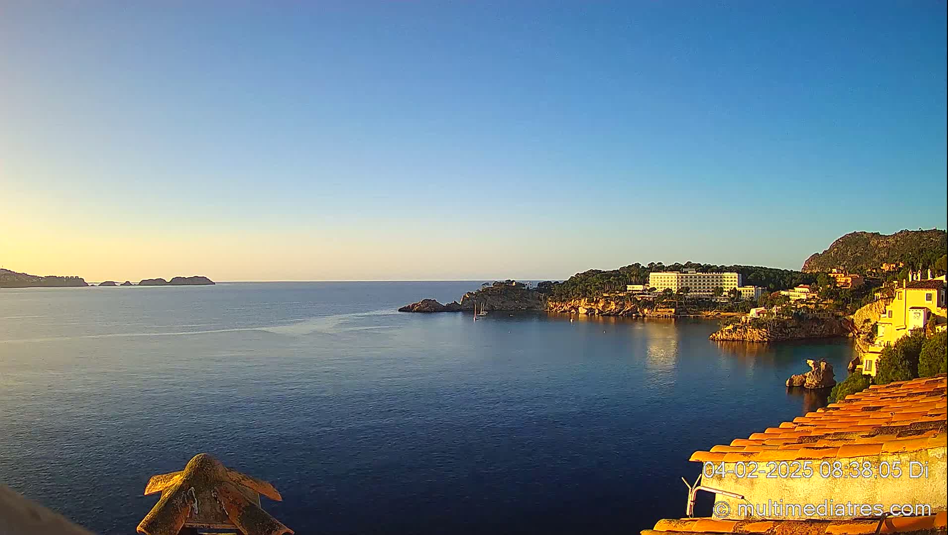 A calm, clear blue sea is seen at sunrise from a high vantage point, overlooking a coastline with buildings and rocky cliffs.