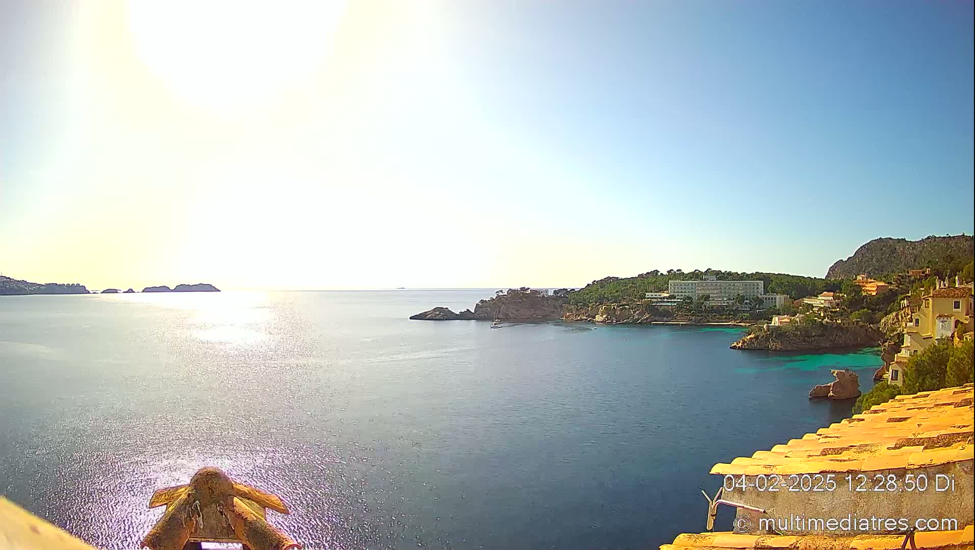 A bright, sunny day reveals a coastal view of a calm sea with rocky outcrops, a hotel, and buildings nestled along the shoreline.