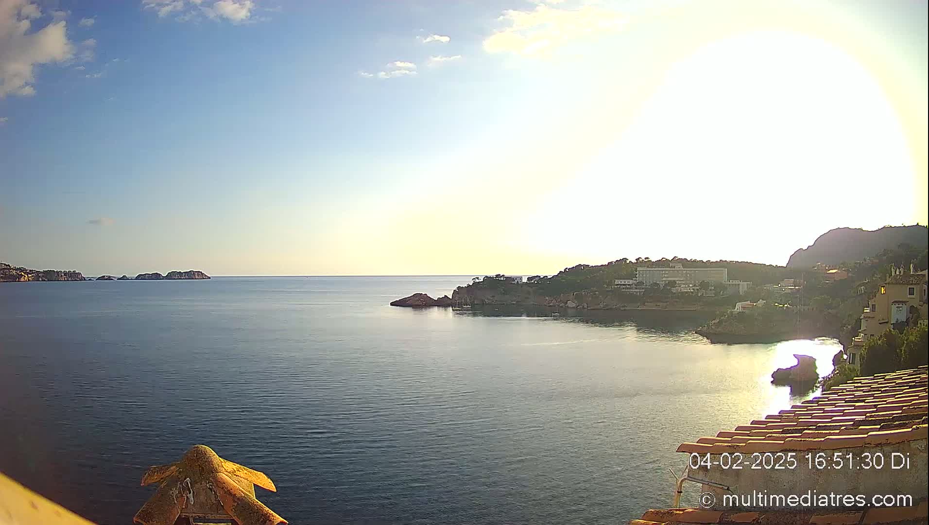 A calm, sunlit sea view shows a coastline with buildings, some small islands in the distance, and a partly cloudy sky.