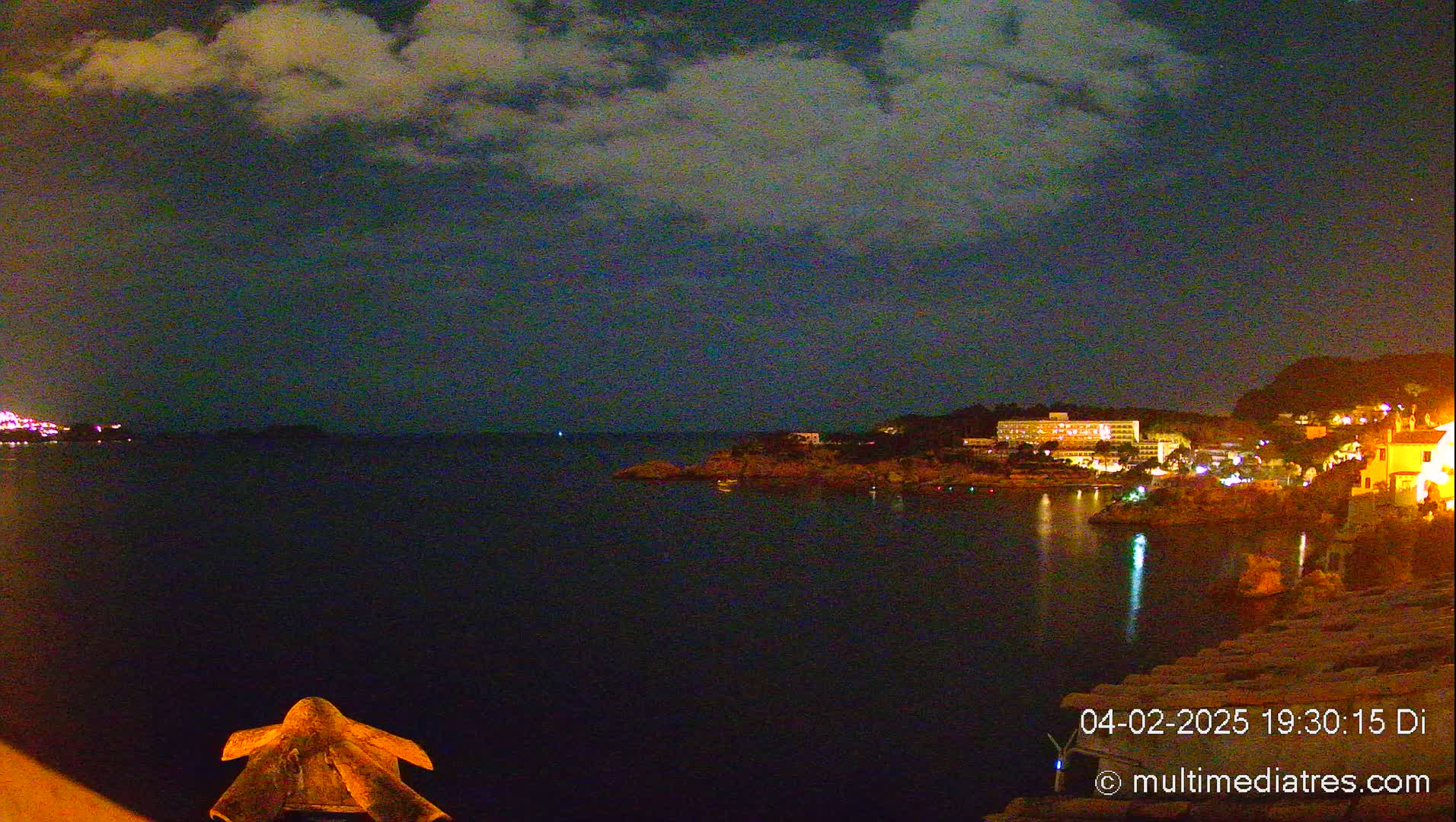 A nighttime view of a calm, dark body of water with illuminated buildings on the opposite shore under a partly cloudy sky.