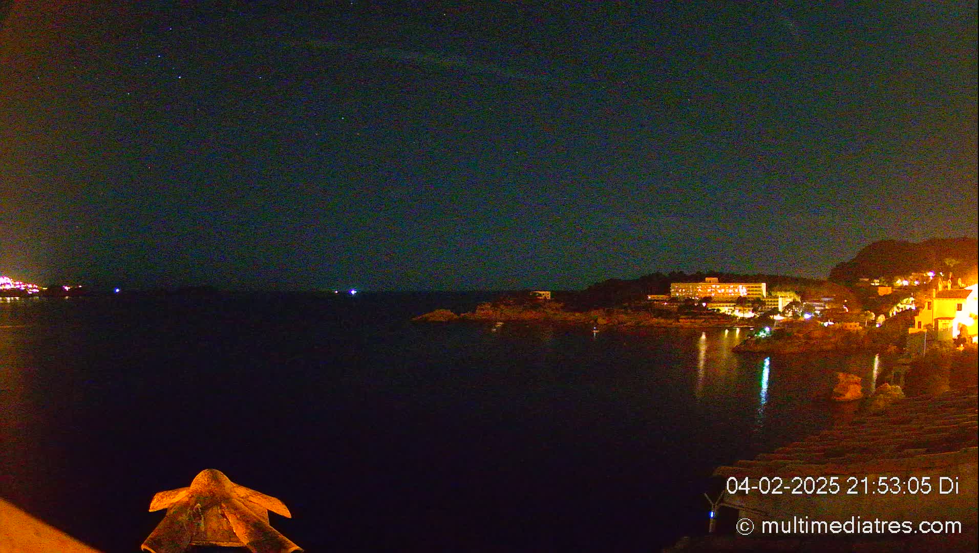 A nighttime view of a dark body of water with a few lights visible on the opposite shore under a clear, starry sky.