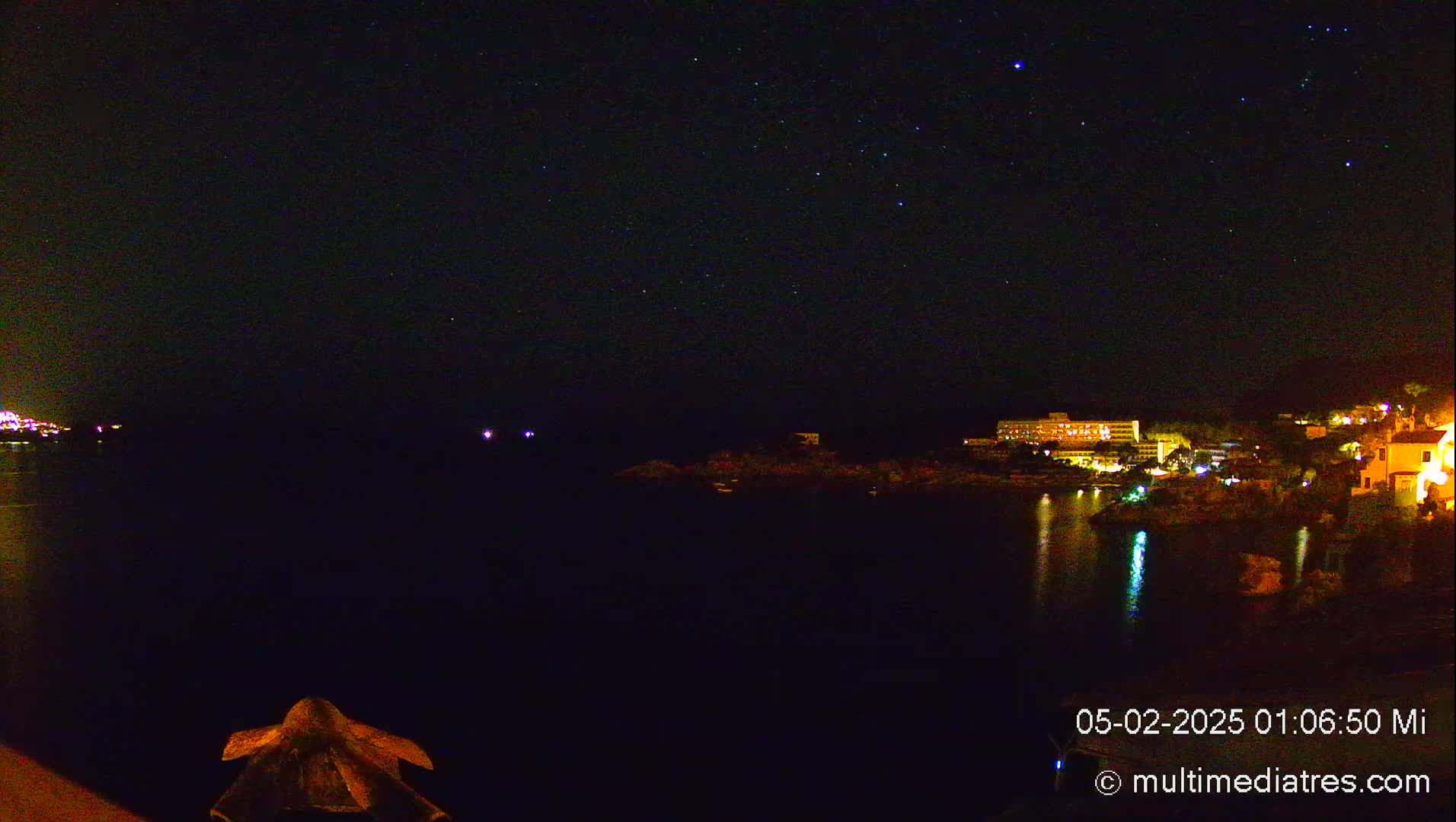 A nighttime view of a dark body of water reflecting the lights of buildings on a distant shoreline under a clear, starry sky.
