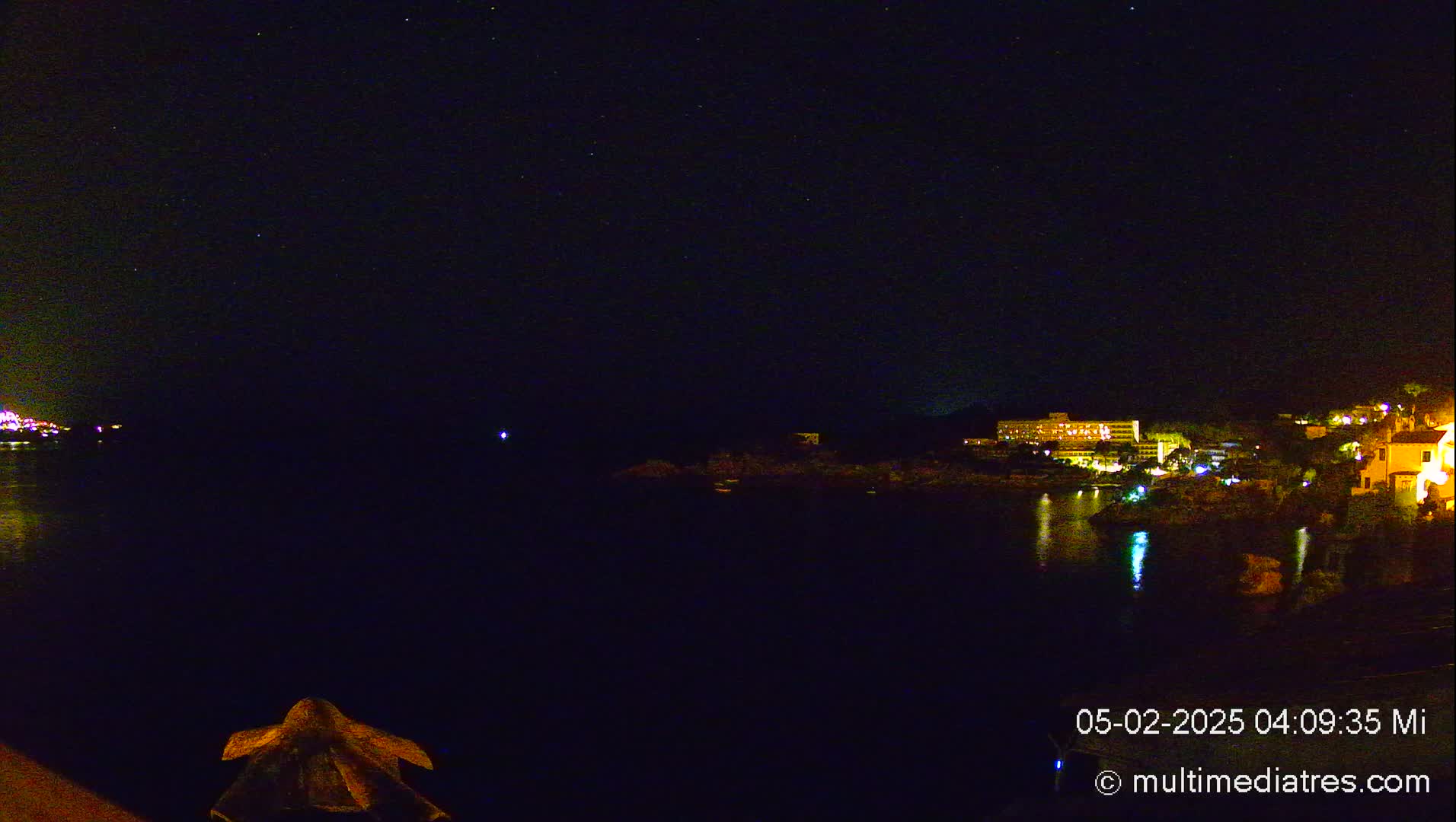 A nighttime view of a dark body of water reflecting the lights of a coastal town under a clear, starry sky.