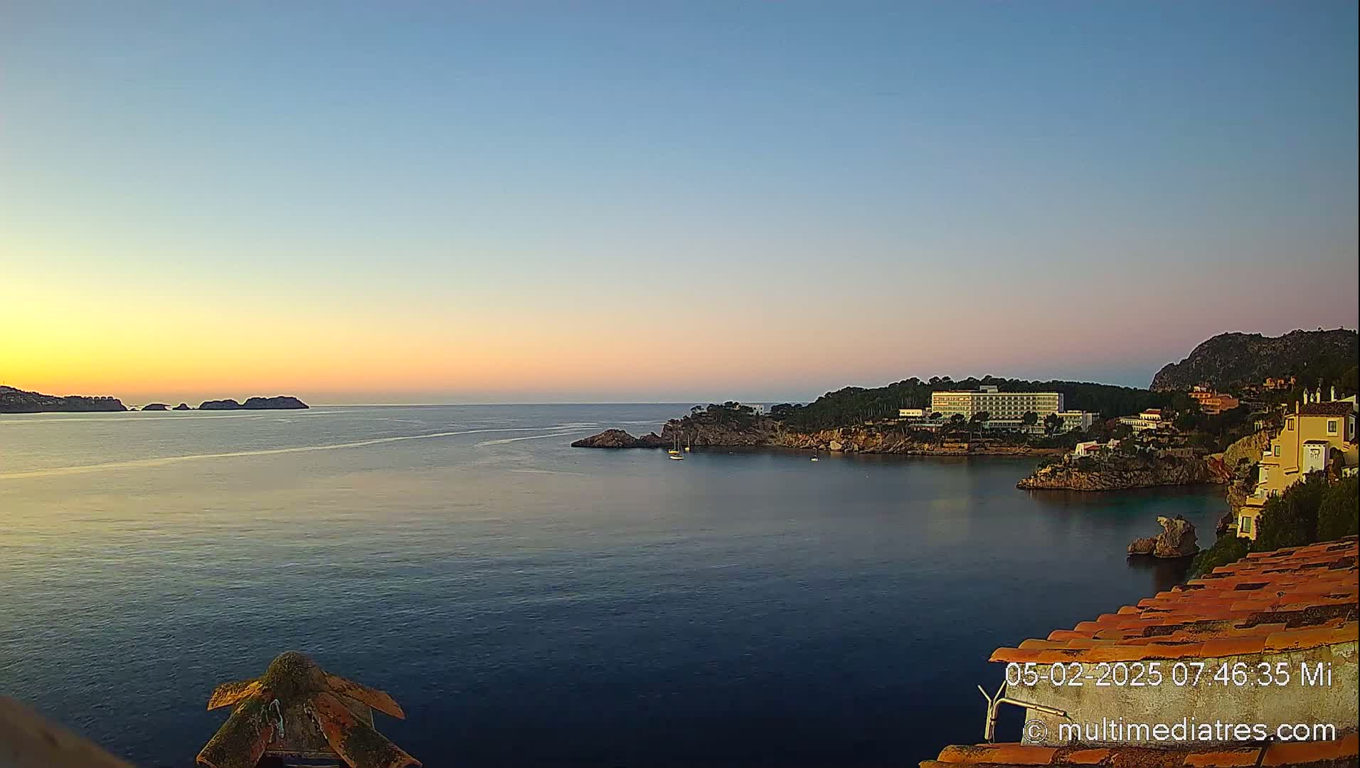 A tranquil sunrise illuminates a calm bay with a hotel and buildings nestled along a rocky coastline, small boats on the water.