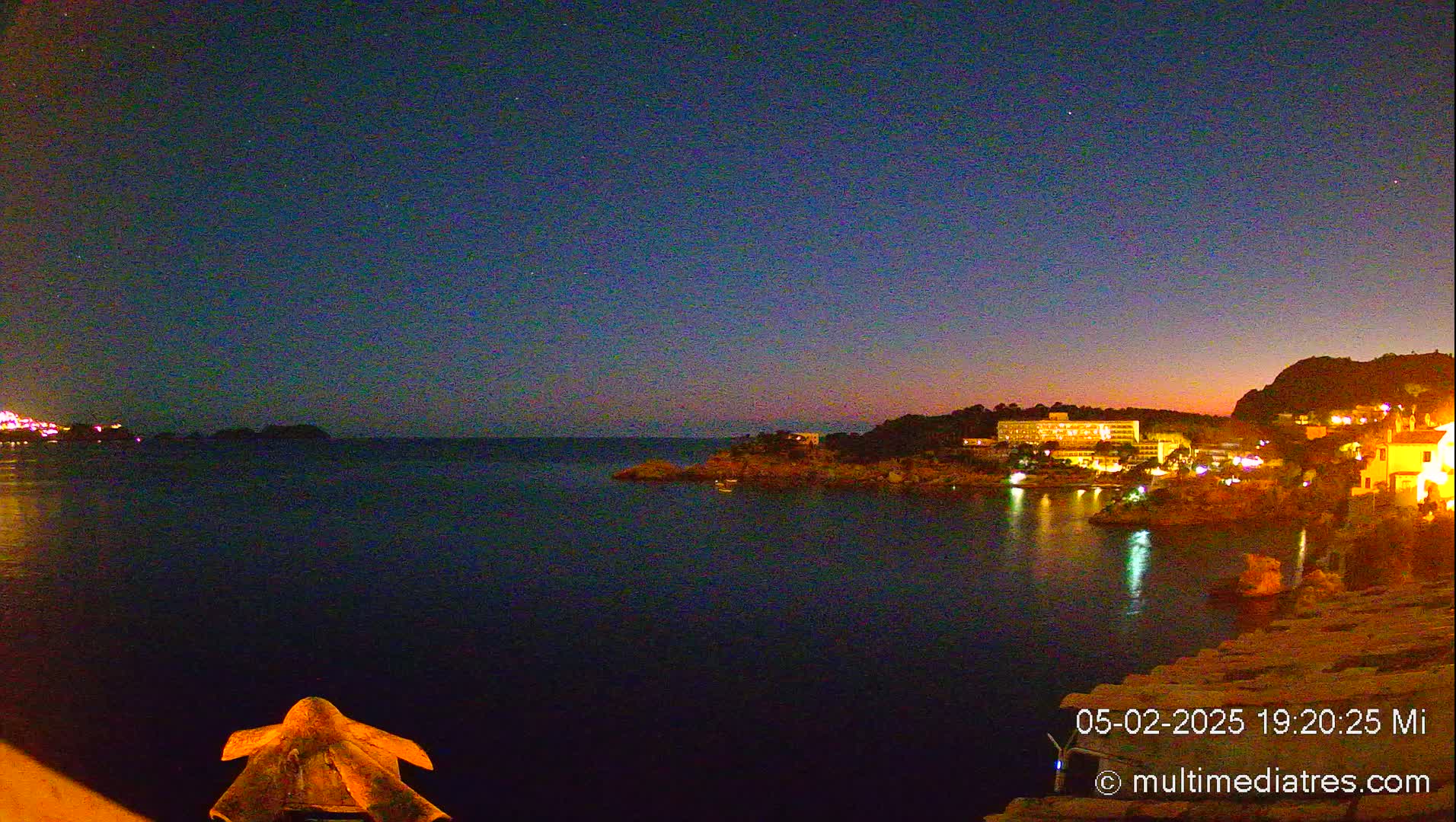 A nighttime view of a calm, dark-blue sea with a coastal town illuminated by lights under a twilight sky.