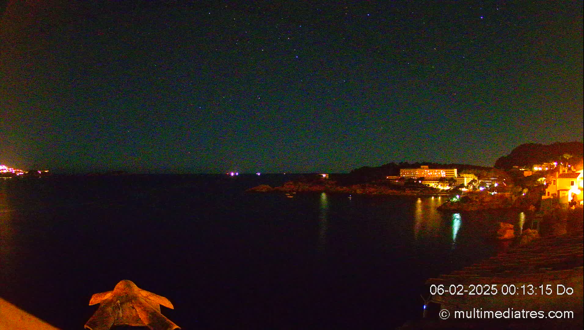 A night scene shows a dark body of water with a building and lights on the opposite shore under a clear, starry night sky.