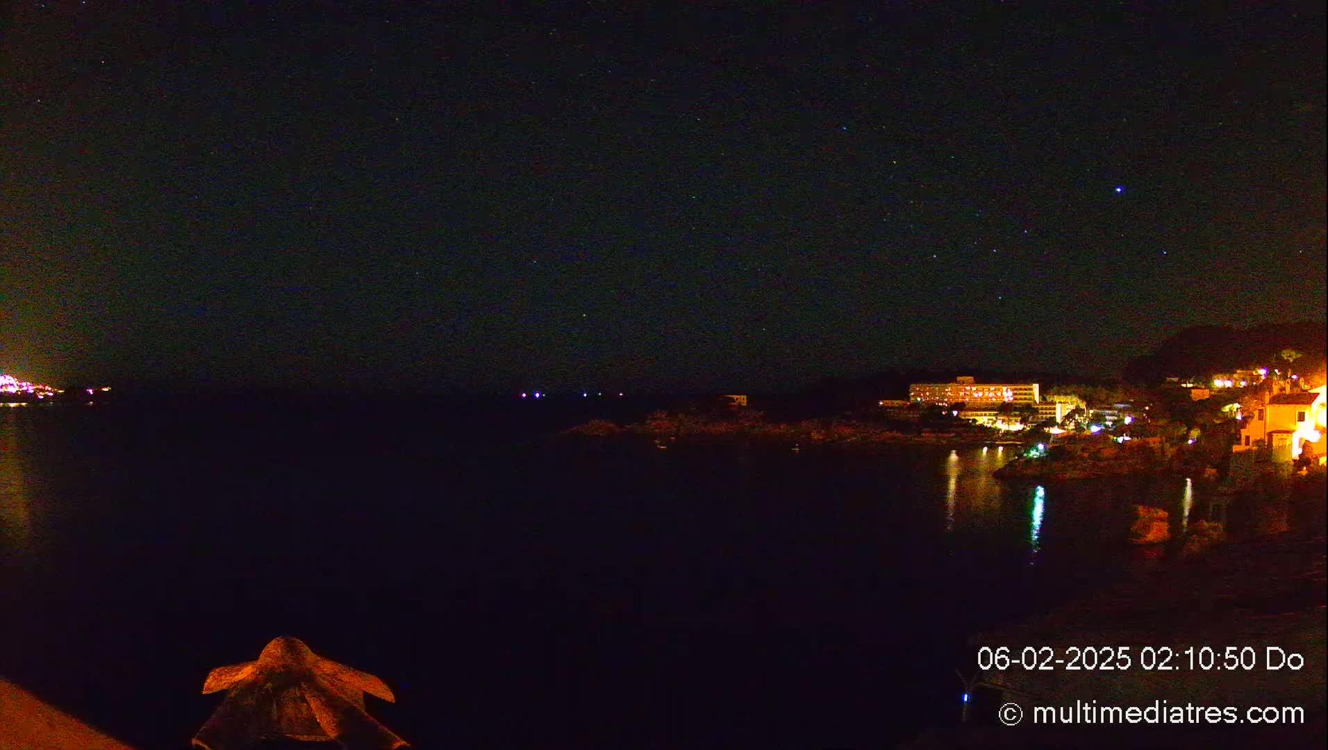 A nighttime view of a dark body of water with a town's lights reflecting on the surface, under a clear, starlit sky.