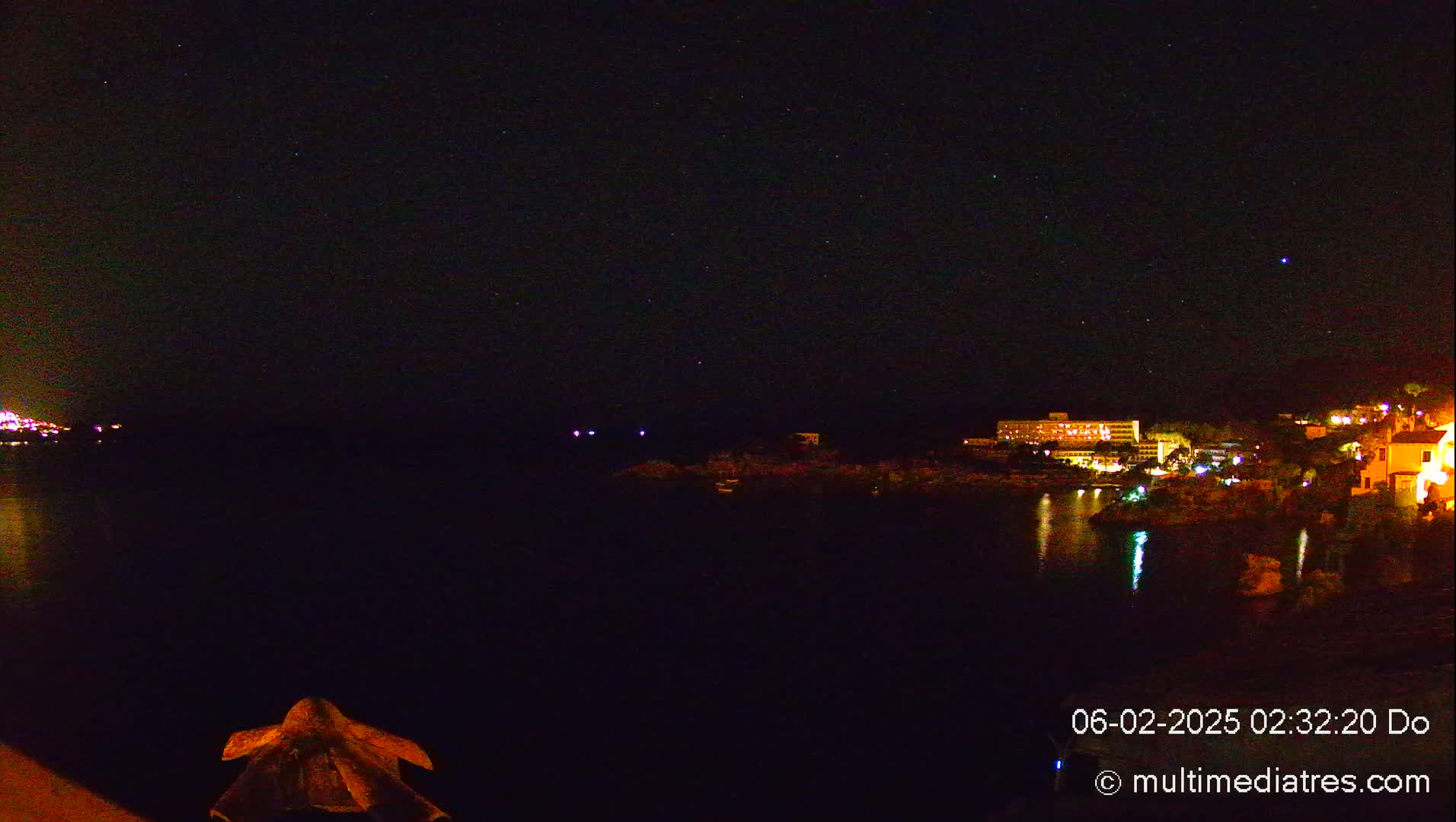 A nighttime view of a dark body of water reflecting lights from buildings on the opposite shore under a clear, starry sky.