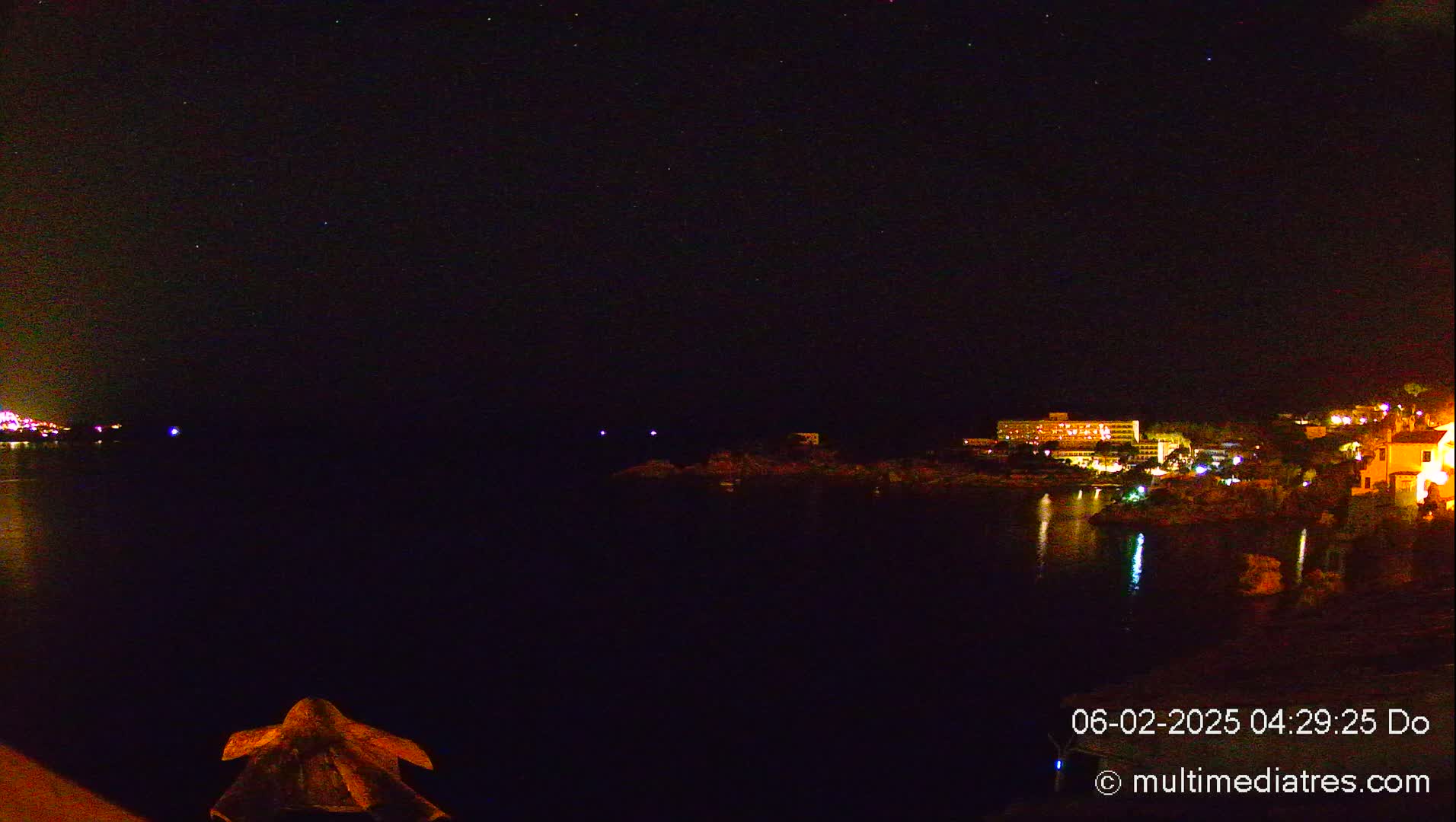 A nighttime view of a calm body of water with a brightly lit shoreline and a starry sky above.