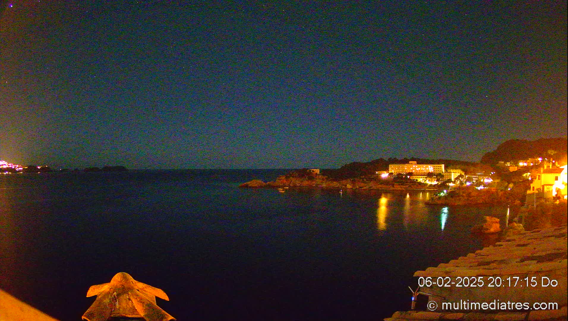 A nighttime view of a calm, dark body of water reflecting the lights of buildings on a hillside under a clear, starry sky.