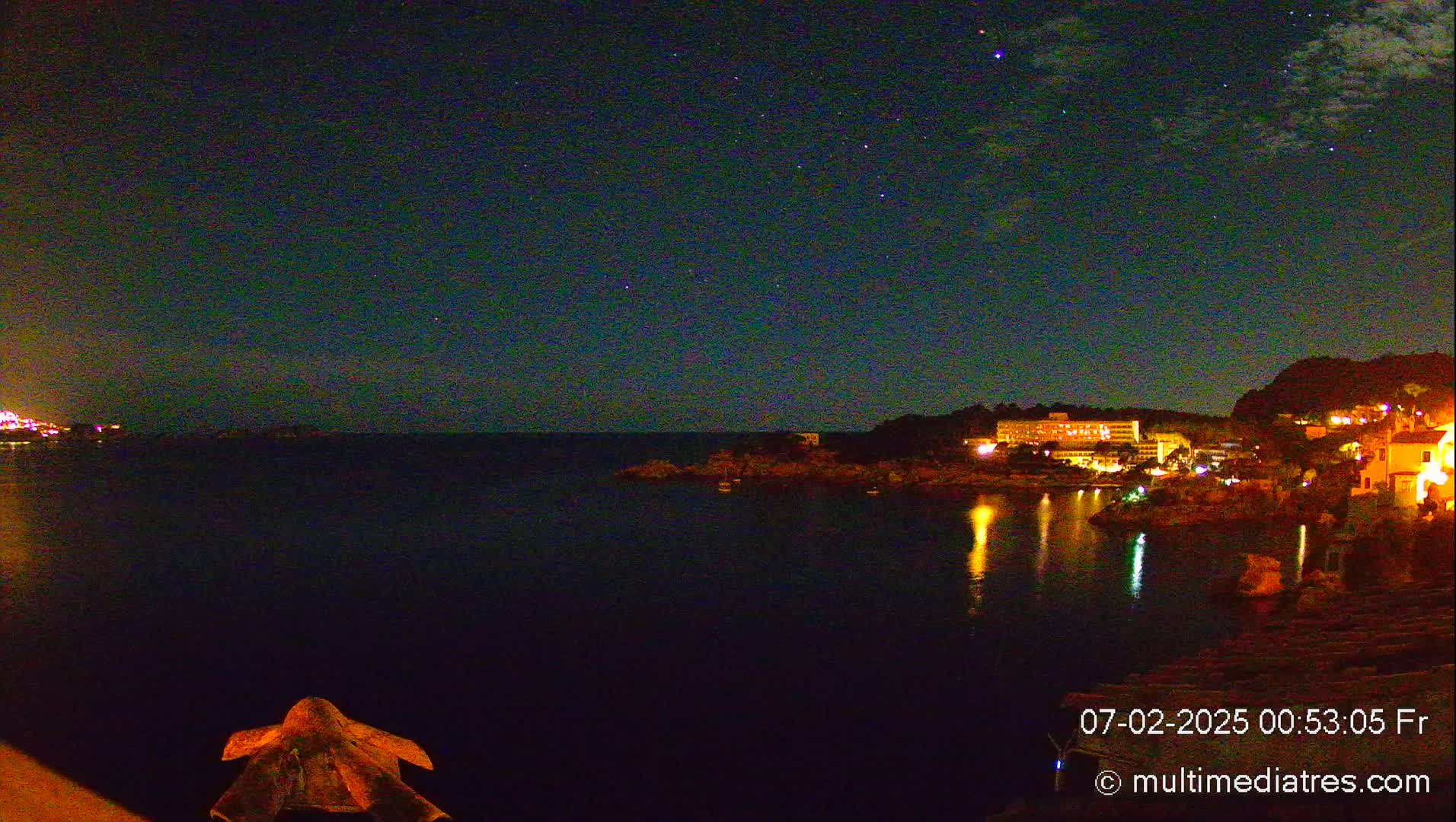 A nighttime view of a calm, dark body of water reflecting the lights of buildings on a rocky shore under a starry sky.