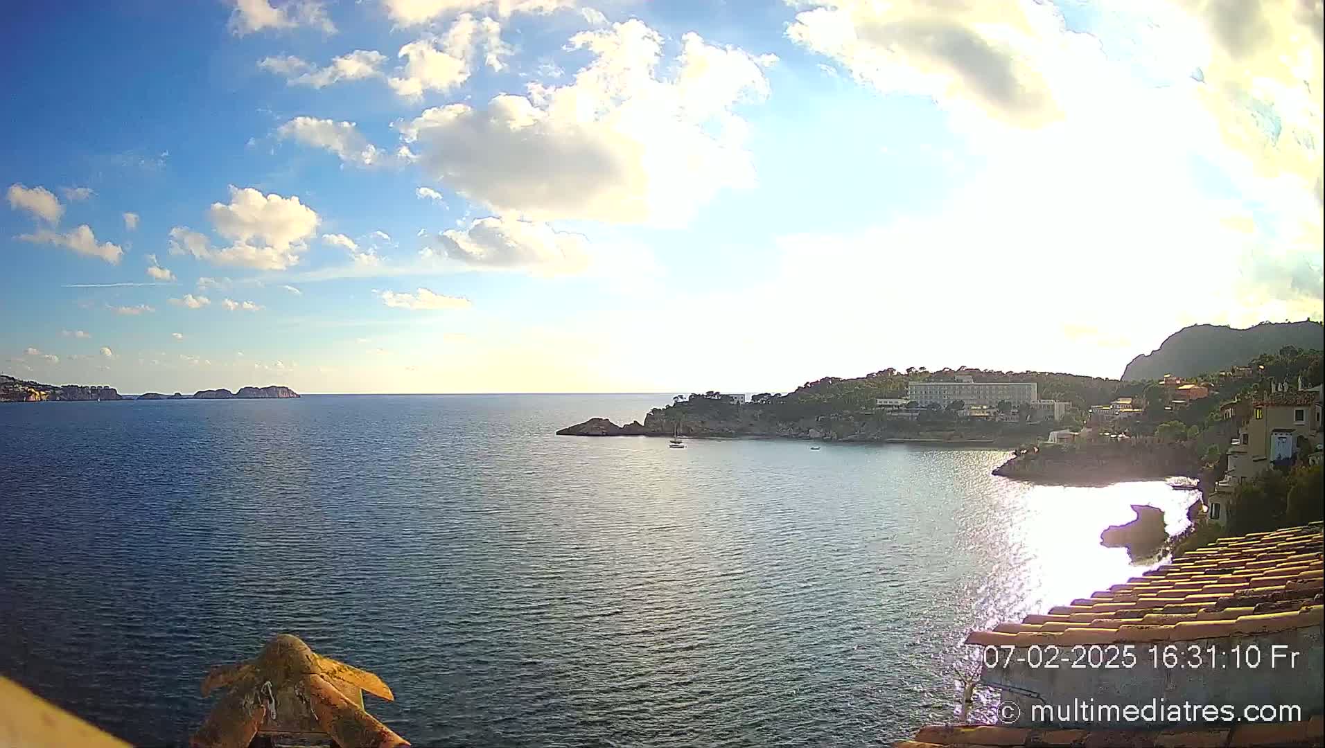 A partly sunny day shows a calm sea with a coastal town and rocky outcroppings under a partly cloudy sky.