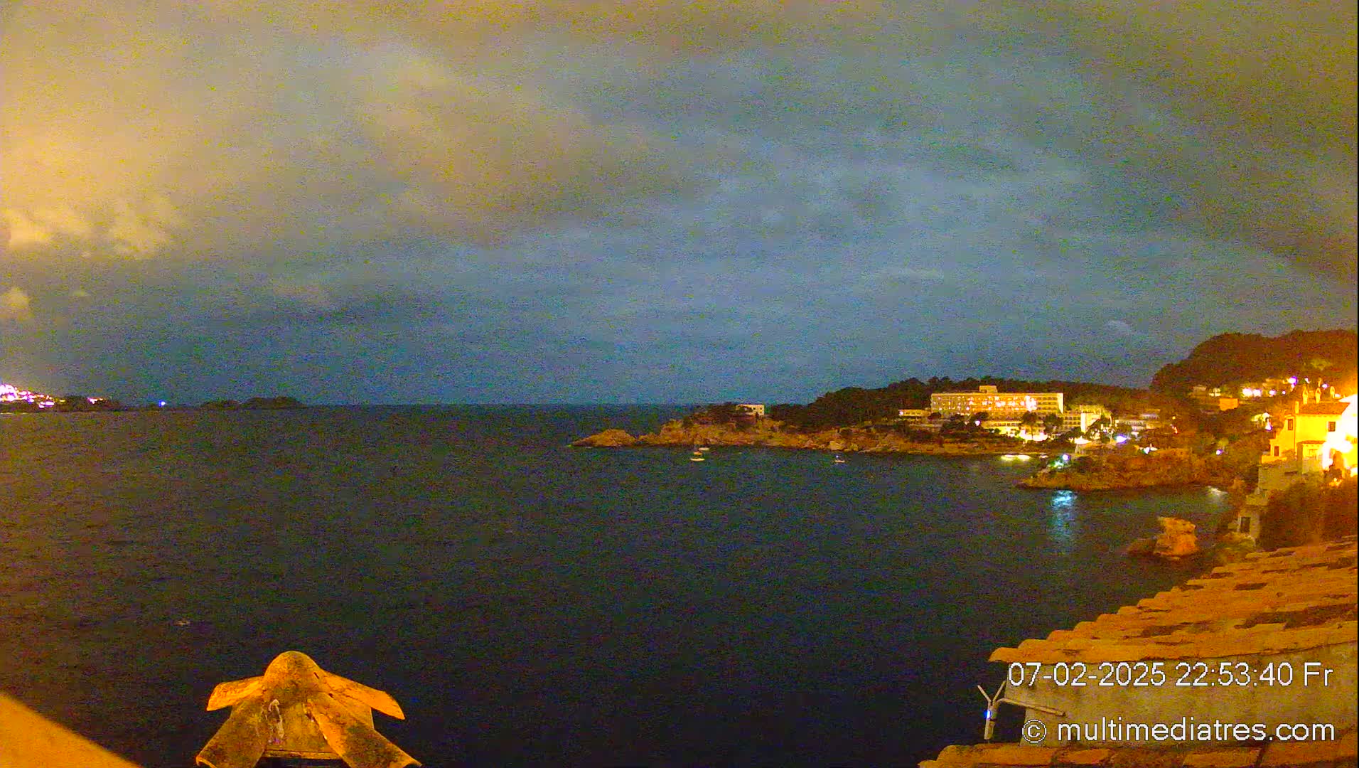 A nighttime coastal scene shows a dark body of water under a cloudy sky, with illuminated buildings and vegetation along the shoreline.