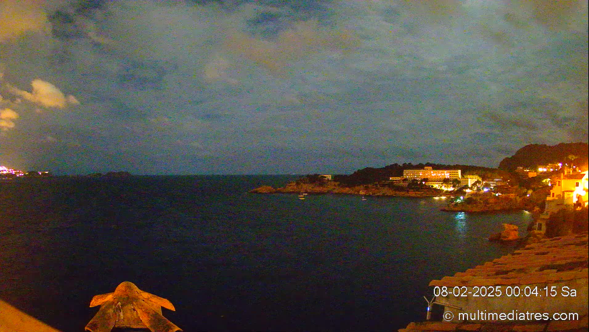 A nighttime view of a calm, dark-blue ocean with a coastline of buildings and trees under a cloudy sky.